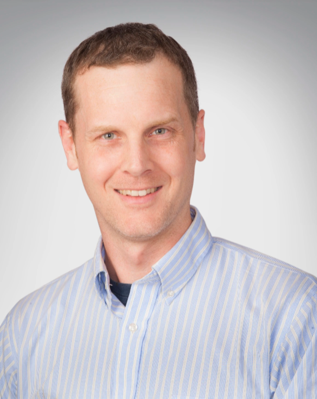 A smiling man with short brown hair wearing a blue and white striped dress shirt against a light gray background.