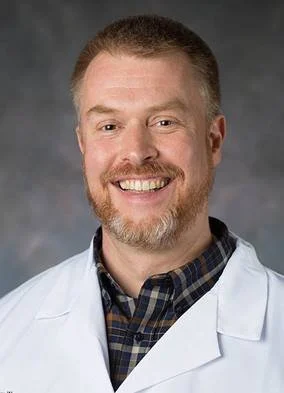 Portrait of a smiling man wearing a white coat over a plaid shirt against a gray background.