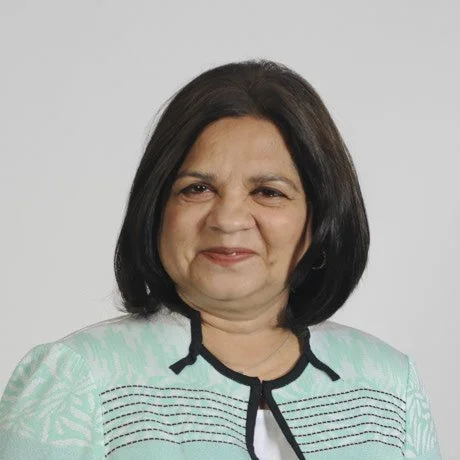 A middle-aged woman with shoulder-length black hair, wearing a light teal jacket with black trim, standing against a plain gray background.
