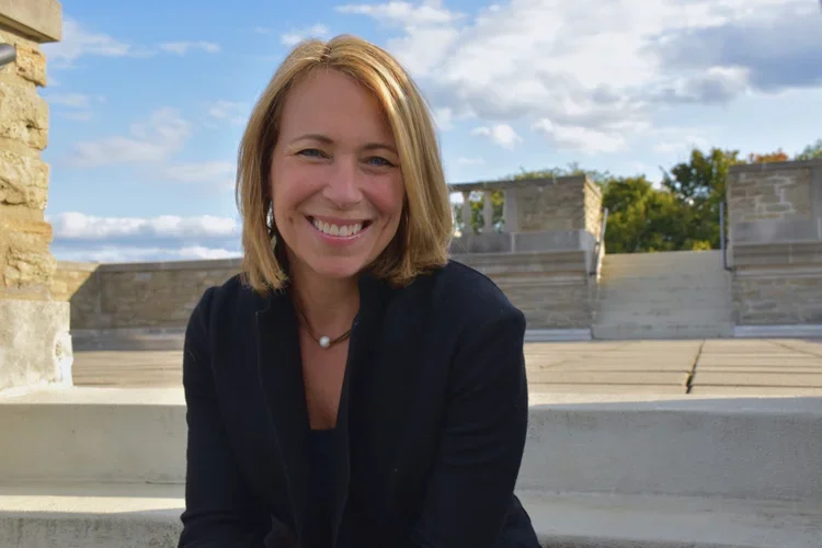 A smiling woman with shoulder-length blonde hair, wearing a black blazer, sitting outdoors on stone steps with a partly cloudy blue sky in the background.