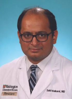 Portrait of a male doctor wearing glasses, a white coat, and a striped tie, against a blue background.