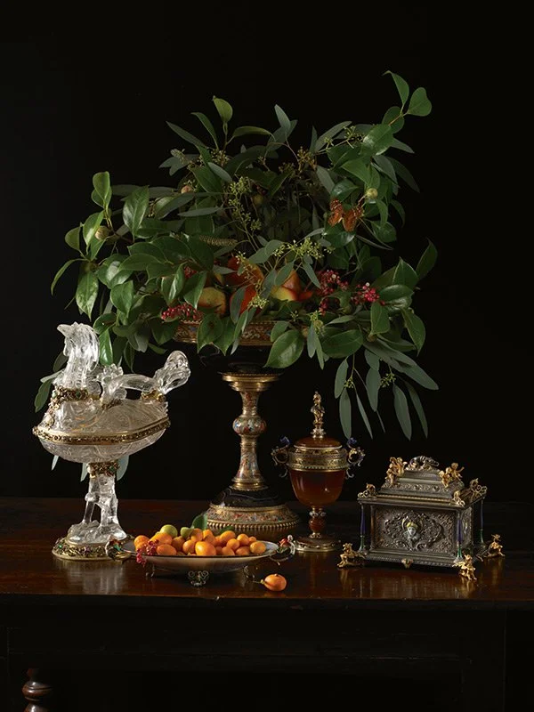 Paulette Tavormina for Sotheby’s — From LUXE: The Art of the Table, a still life in her Old Master style featuring a floral centerpiece with fruits, ornate silver objects on a dark surface, set against a black background.