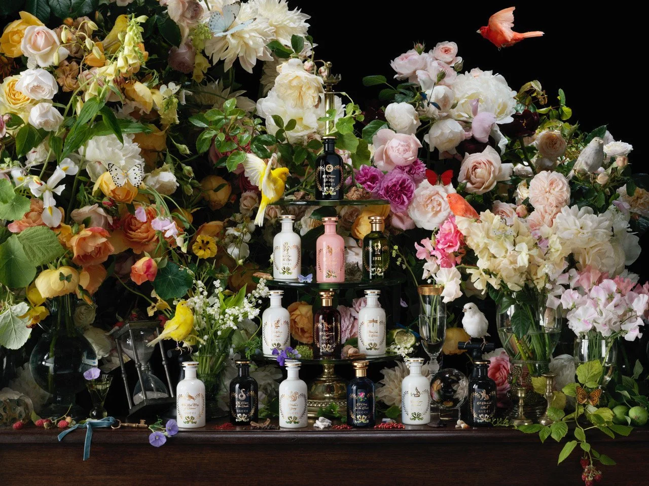 A Paulette Tavormina photograph from Gucci’s The Alchemist’s Garden campaign: a lush floral scene with roses, peonies, birds, and ornate perfume bottles on a dark wooden table