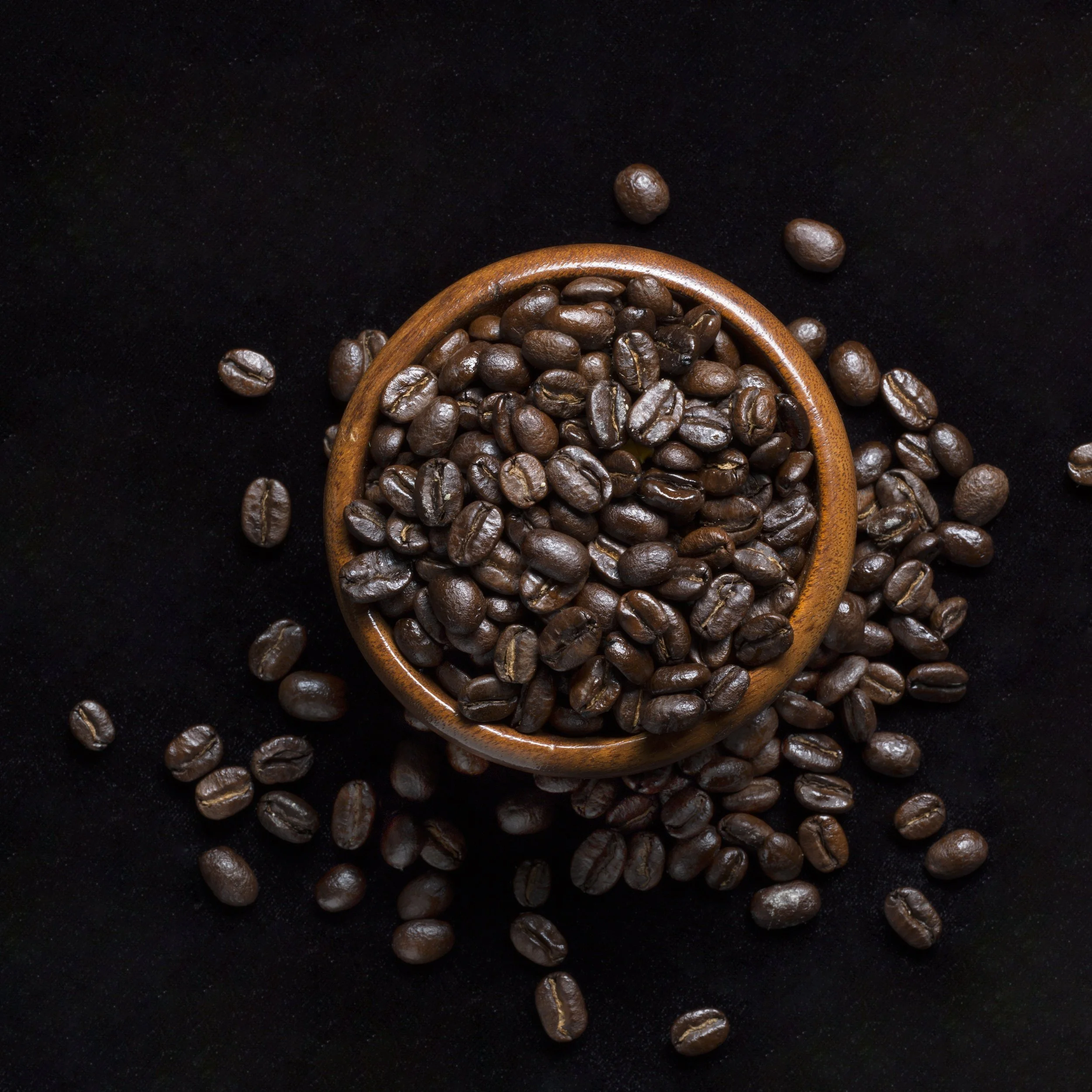 Paulette Tavormina for GOOP: Roasted coffee beans in a wooden bowl on a black surface, with scattered beans around.
