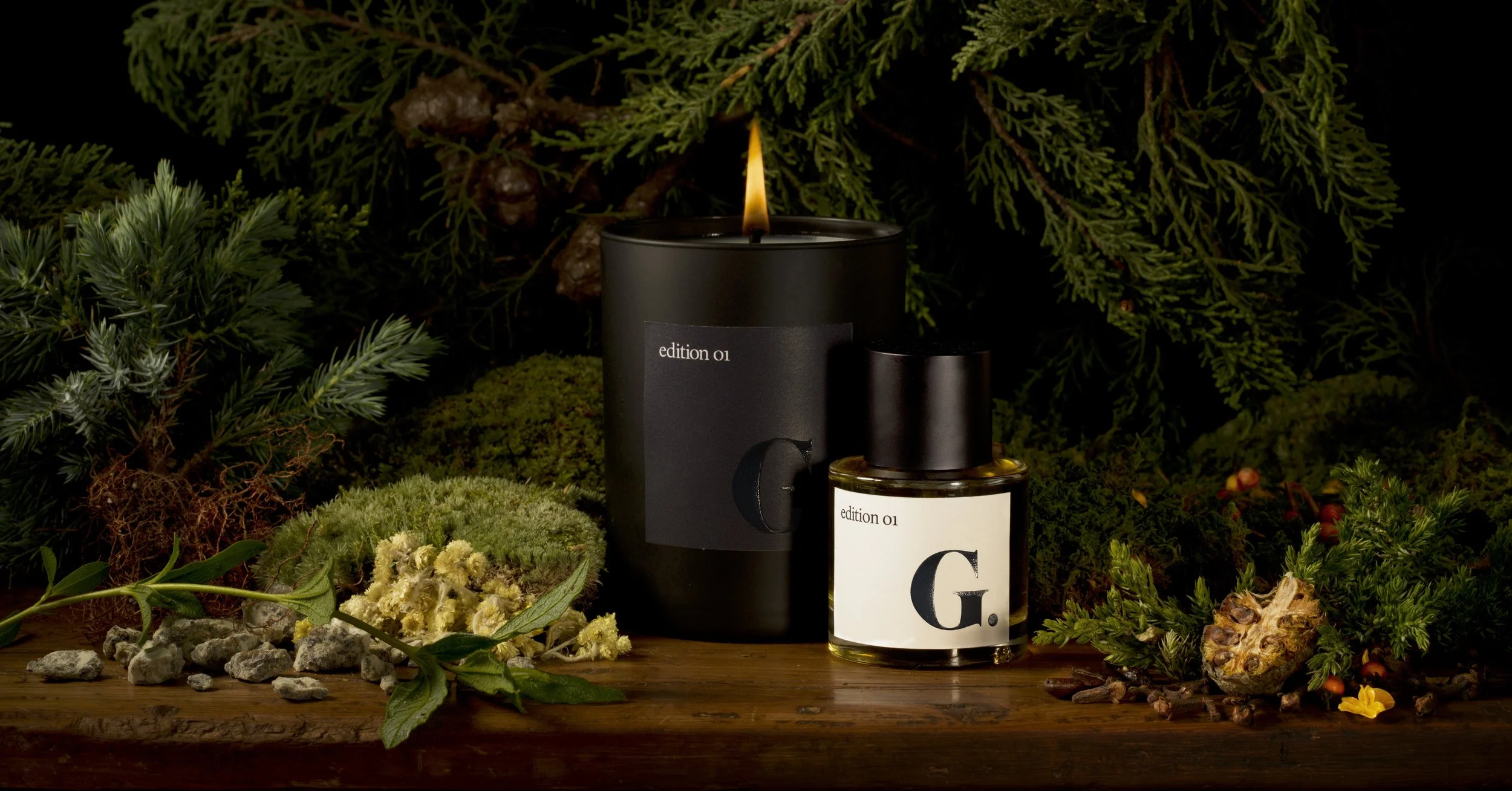 Paulette Tavormina photograph for GOOP’s seasonal perfume: A lit 'Edition 01' candle and perfume bottle set among moss, yellow flowers, and greenery on a wooden surface.