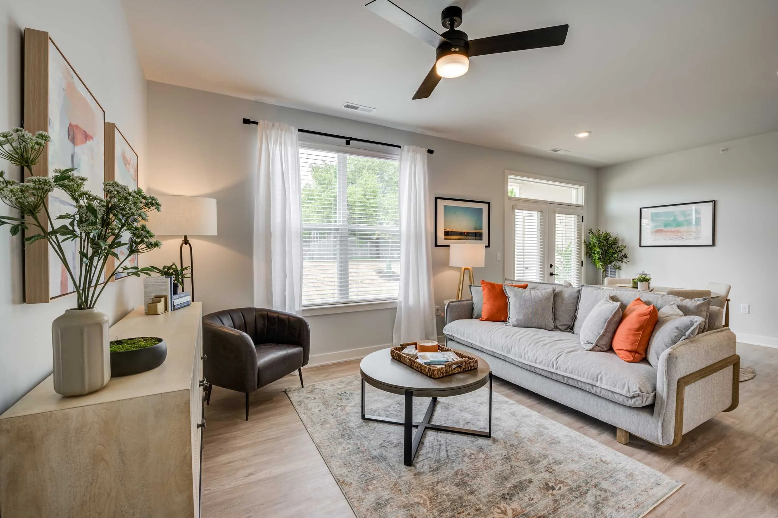 A living room with large window, white curtains, beige sofa with orange pillows, a round coffee table with a tray, a gray rug, a white side table with a lamp and decorative items, framed landscape photos on the wall, a ceiling fan, and a plant.