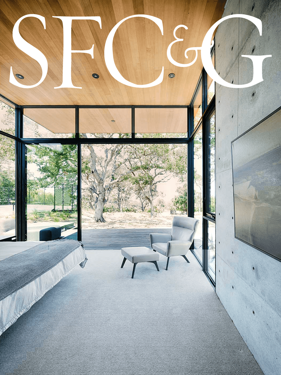 Modern interior with floor-to-ceiling windows, light wood ceiling, concrete wall, and beige armchair with ottoman next to a bed. View of trees and nature outside.
