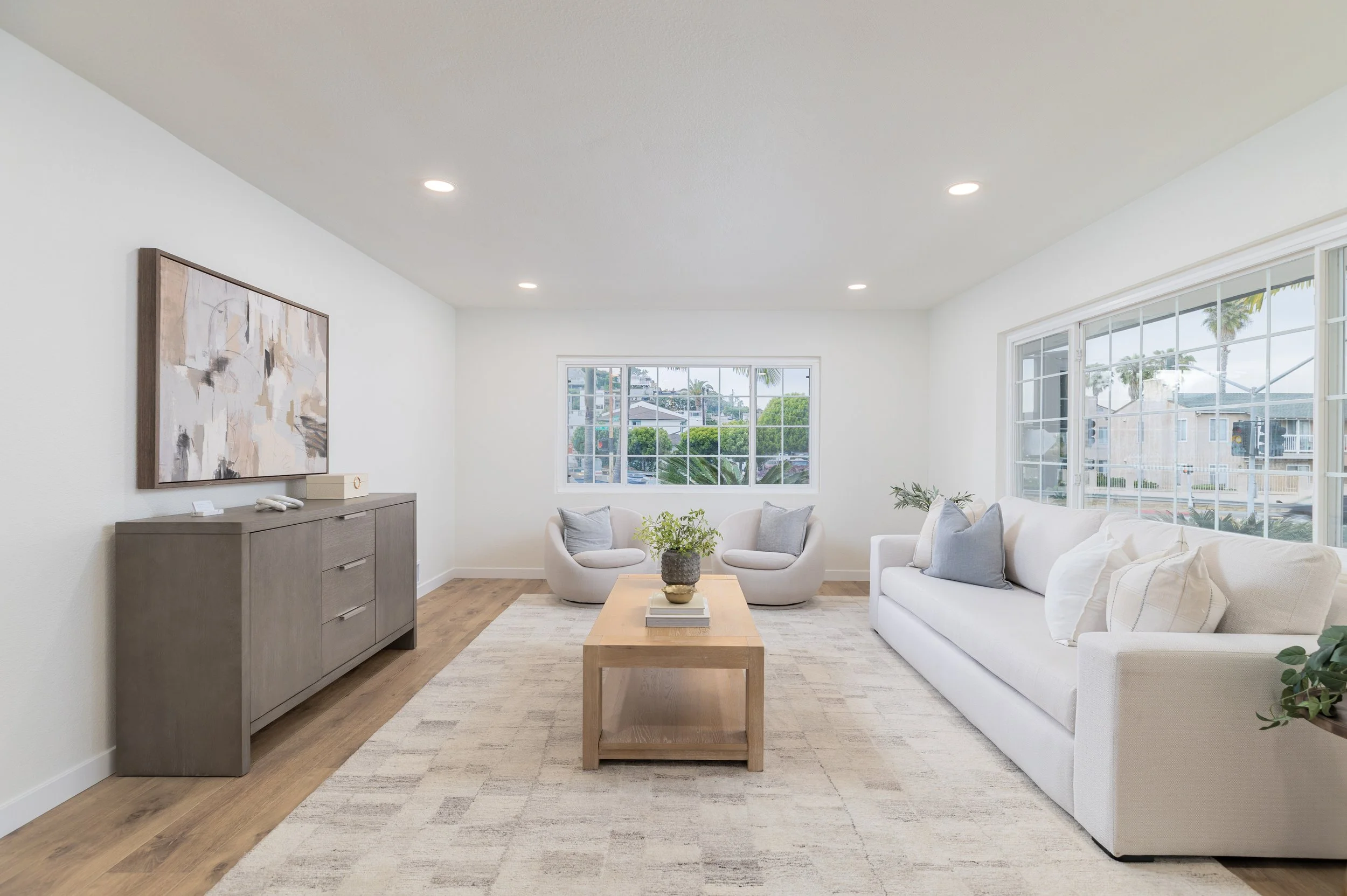 Bright living room with white walls, large windows, white sofa with gray pillows, two white armchairs with gray pillows, a light wood coffee table with a plant and books, a gray sideboard with abstract art above, and a beige area rug.