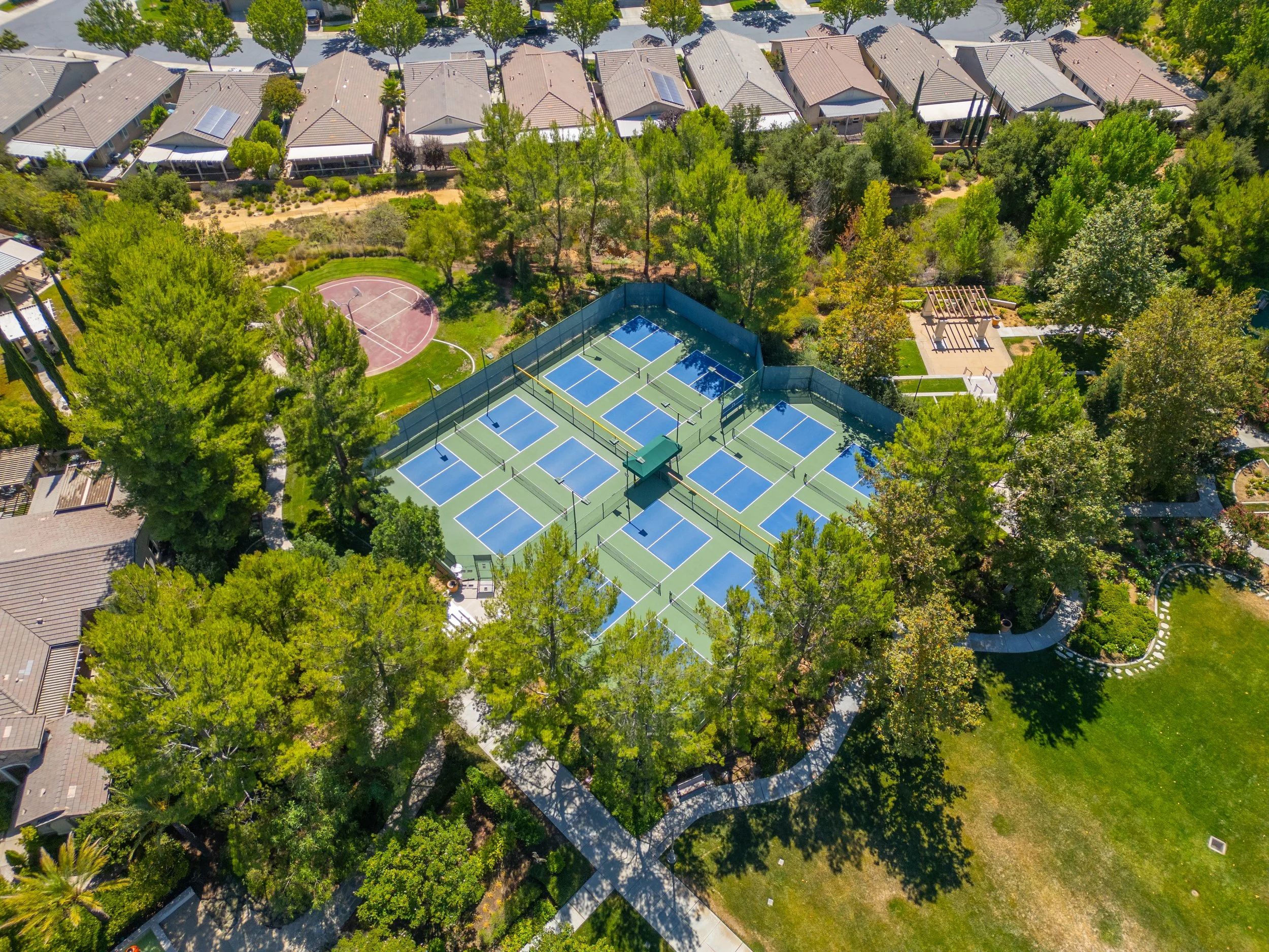 An aerial view of a neighborhood with a tennis court complex, a basketball court, and a playground, surrounded by trees and houses.