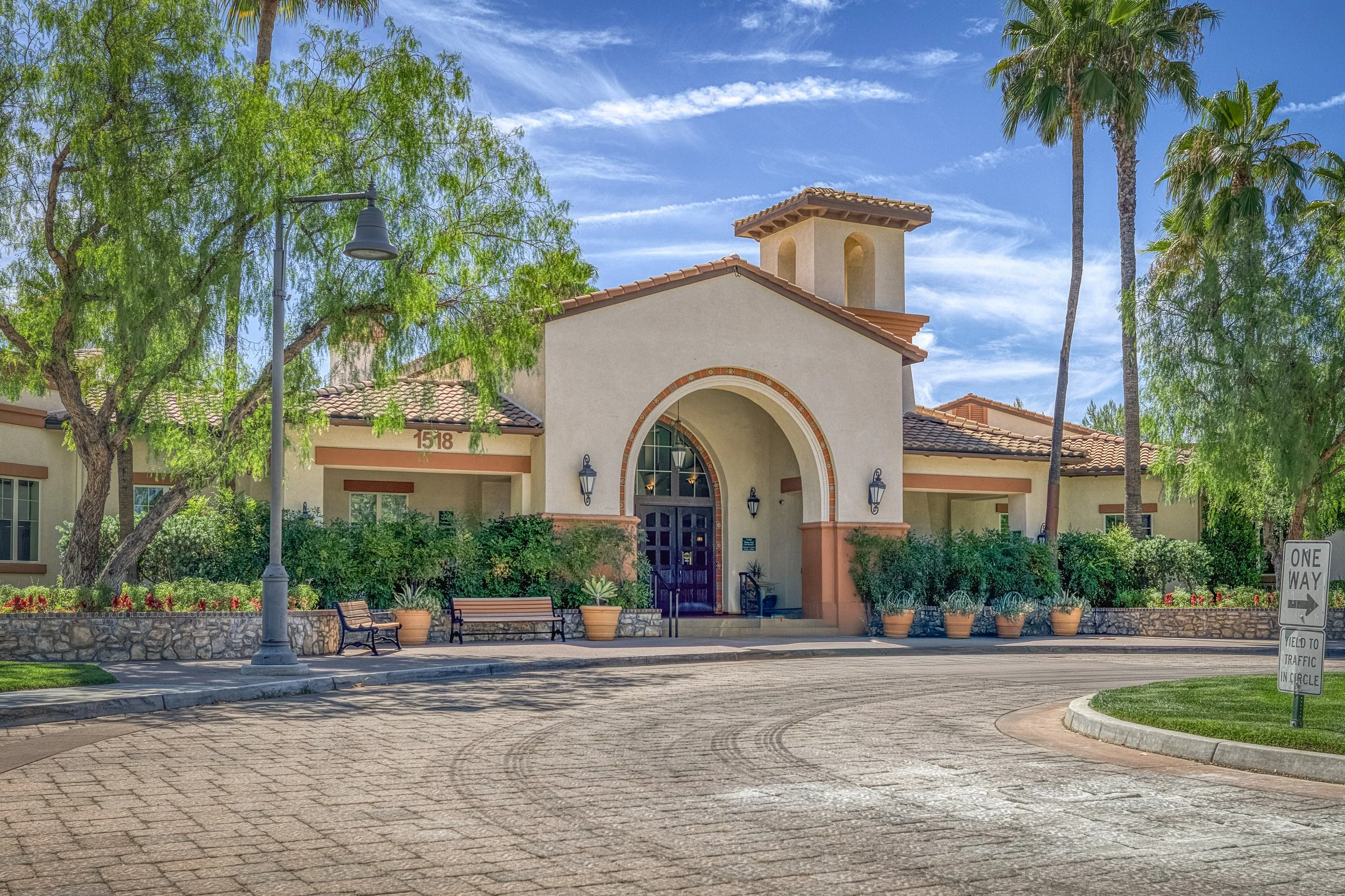 A residential community entrance with a building featuring a large archway, palm trees, a lamppost, benches, potted plants, and a one-way traffic sign under a partly cloudy sky.