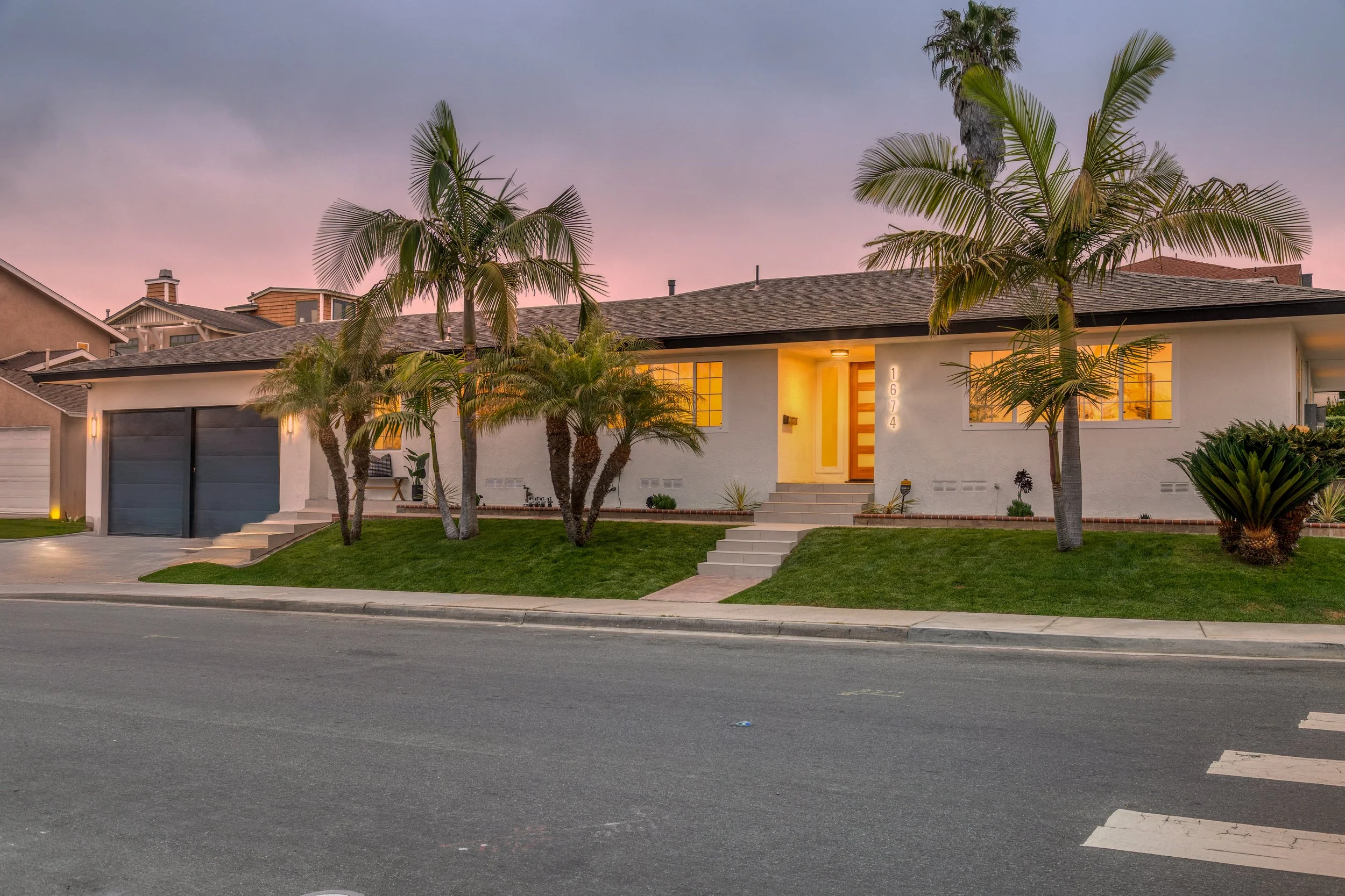A modern single-story house with a white exterior, palm trees in the front yard, and a garage with dark doors. The house is illuminated and has a pathway leading to the front door, with a pink and purple sunset sky in the background.
