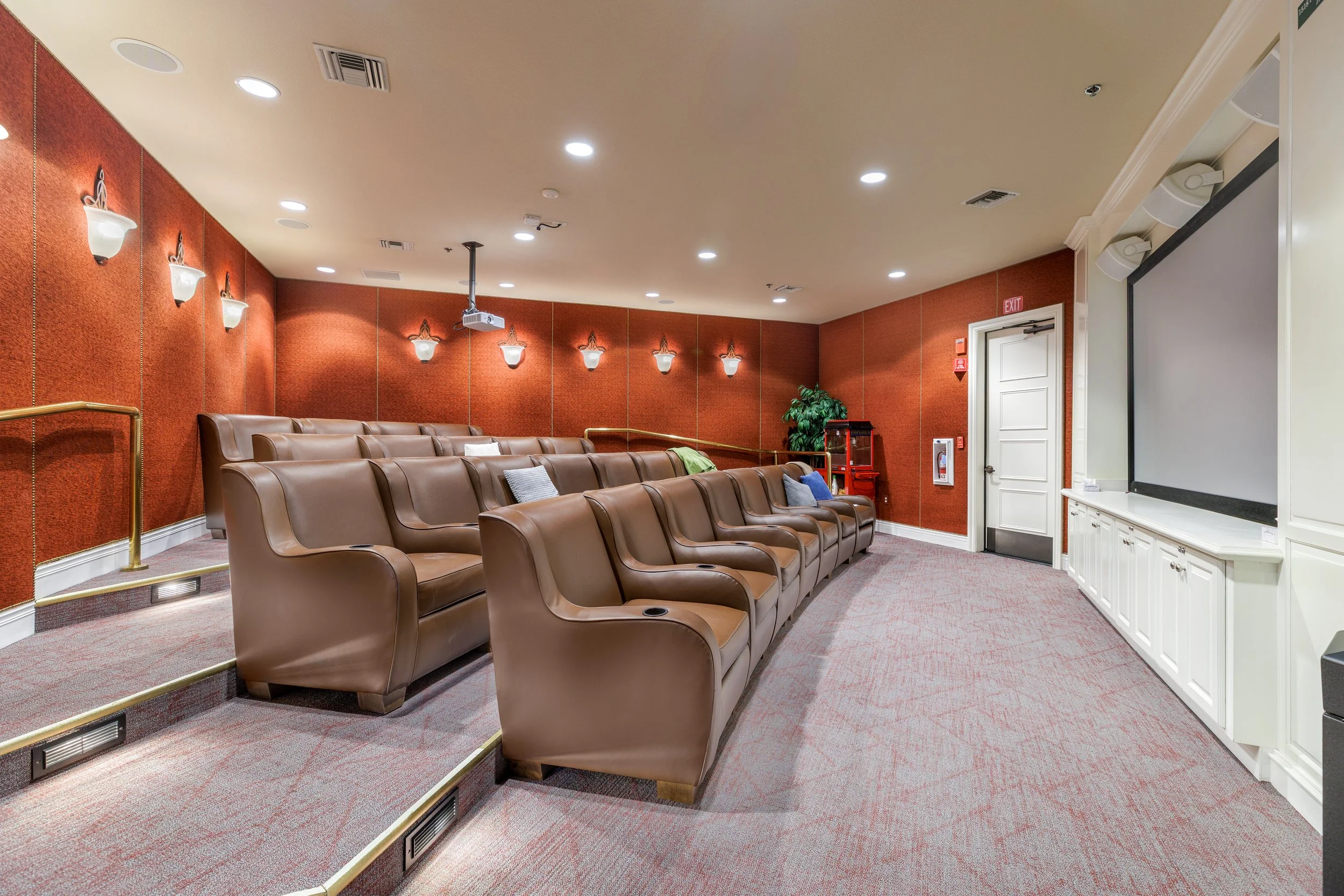 Empty small auditorium with brown leather seats arranged in tiered rows, white and red painted walls, wall-mounted lights, a white door with an exit sign, a projector hanging from the ceiling, a potted plant, and a large projection screen at the fron