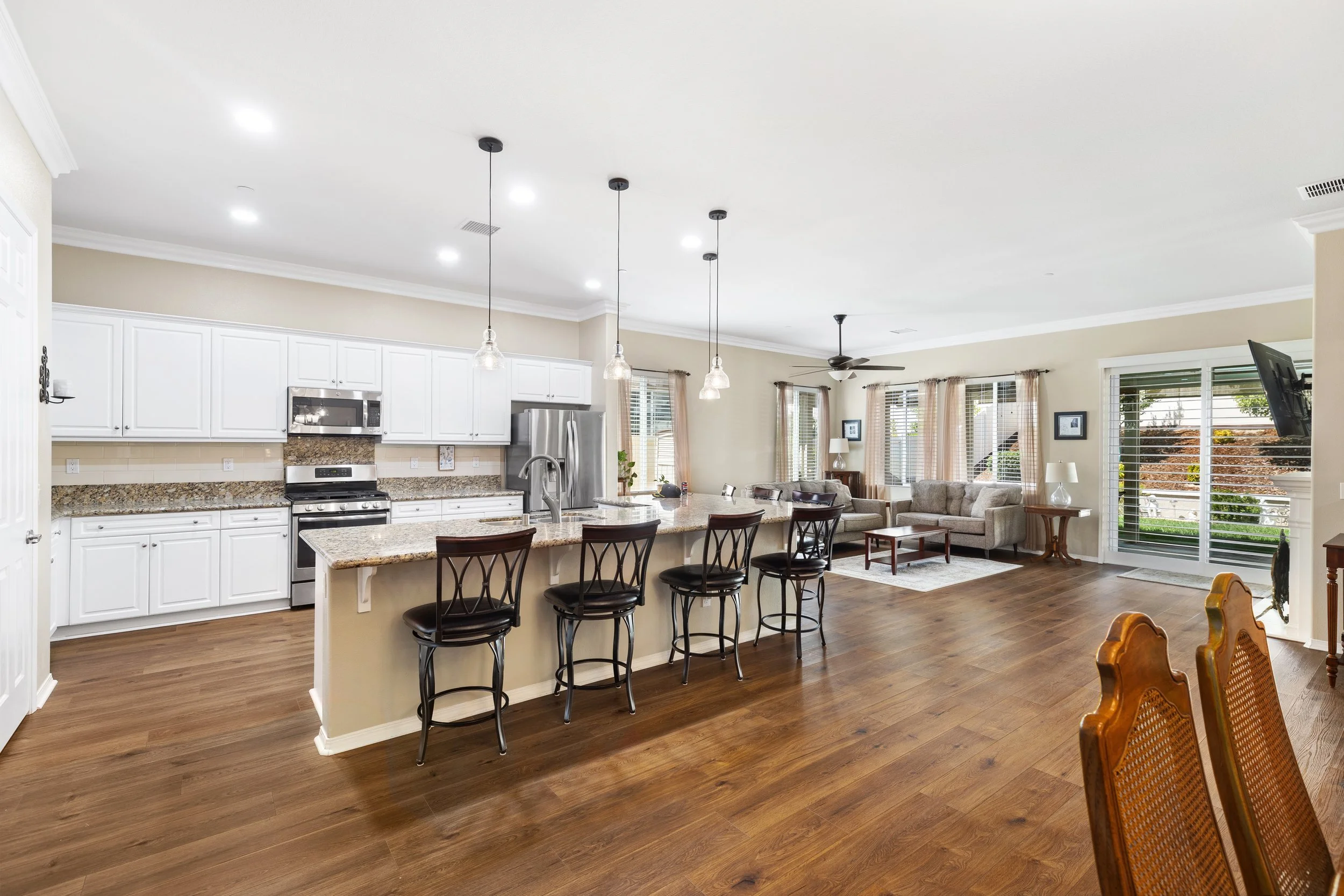 Open-concept kitchen and living room with white cabinets, granite countertops, stainless steel appliances, bar stools at kitchen island, hardwood floors, beige walls, and large windows with curtains.