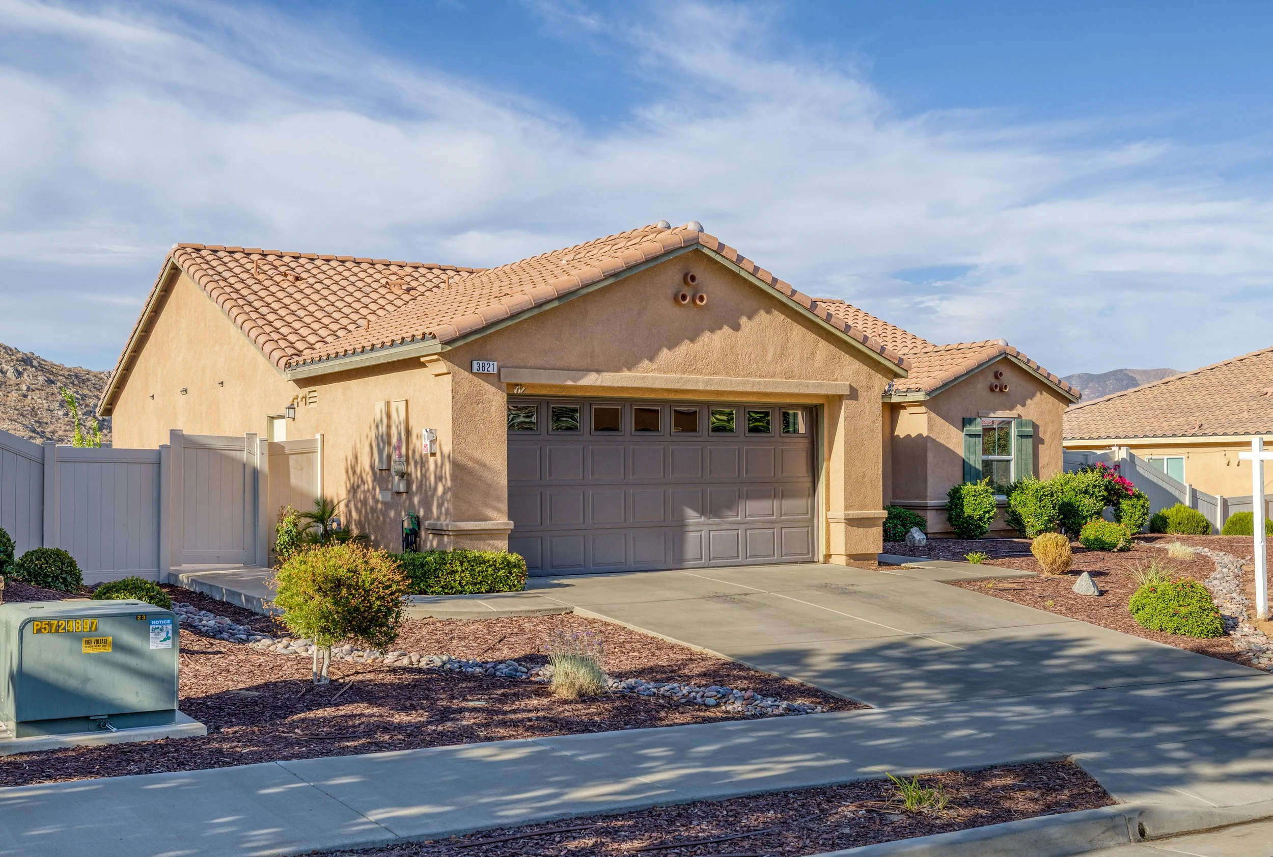 A modern beige house with a brown tiled roof, a closed garage door, a driveway, desert landscaping, shrubs, and a white fence, under a partly cloudy sky.