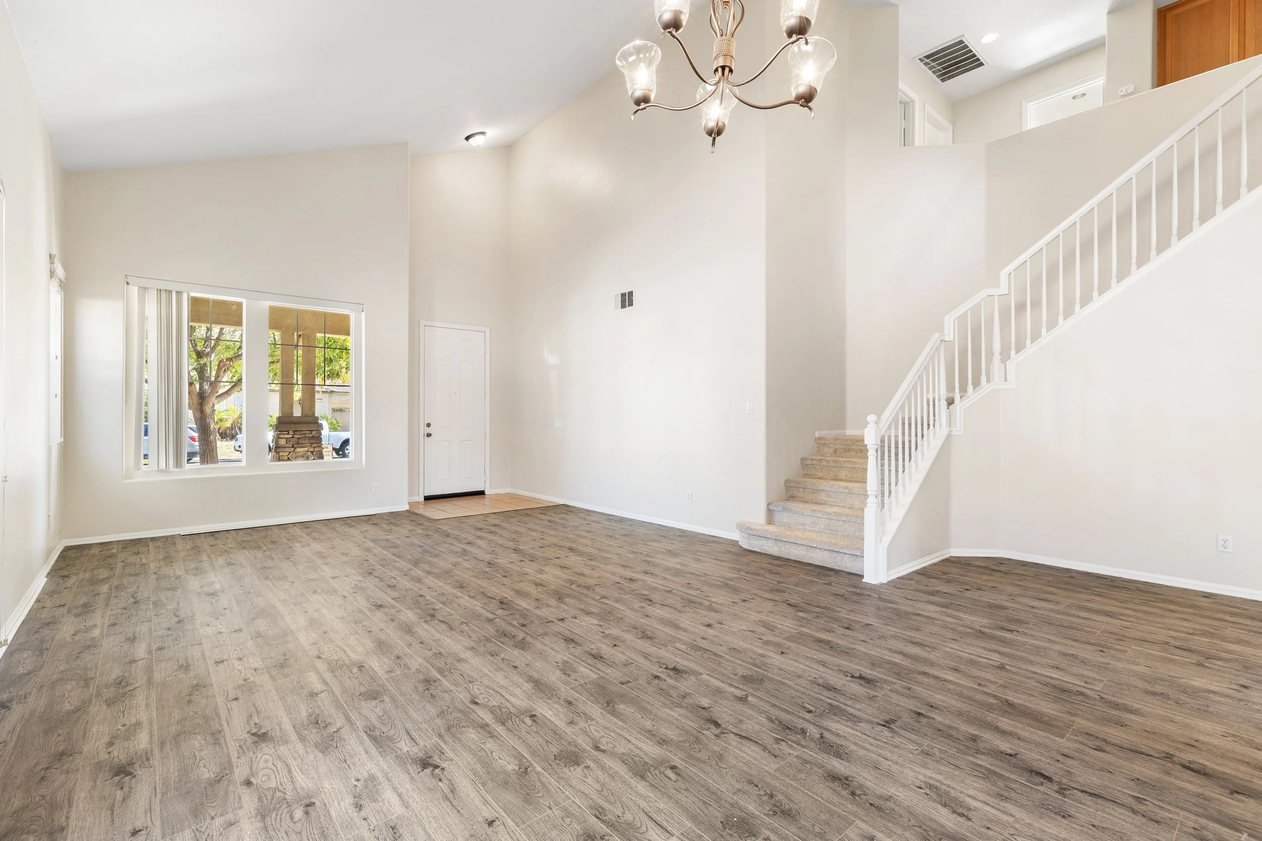 Empty living room with light-colored walls, wood laminate flooring, a chandelier hanging from the ceiling, a window with vertical blinds, a front door, and a staircase with carpeted steps and a white banister.