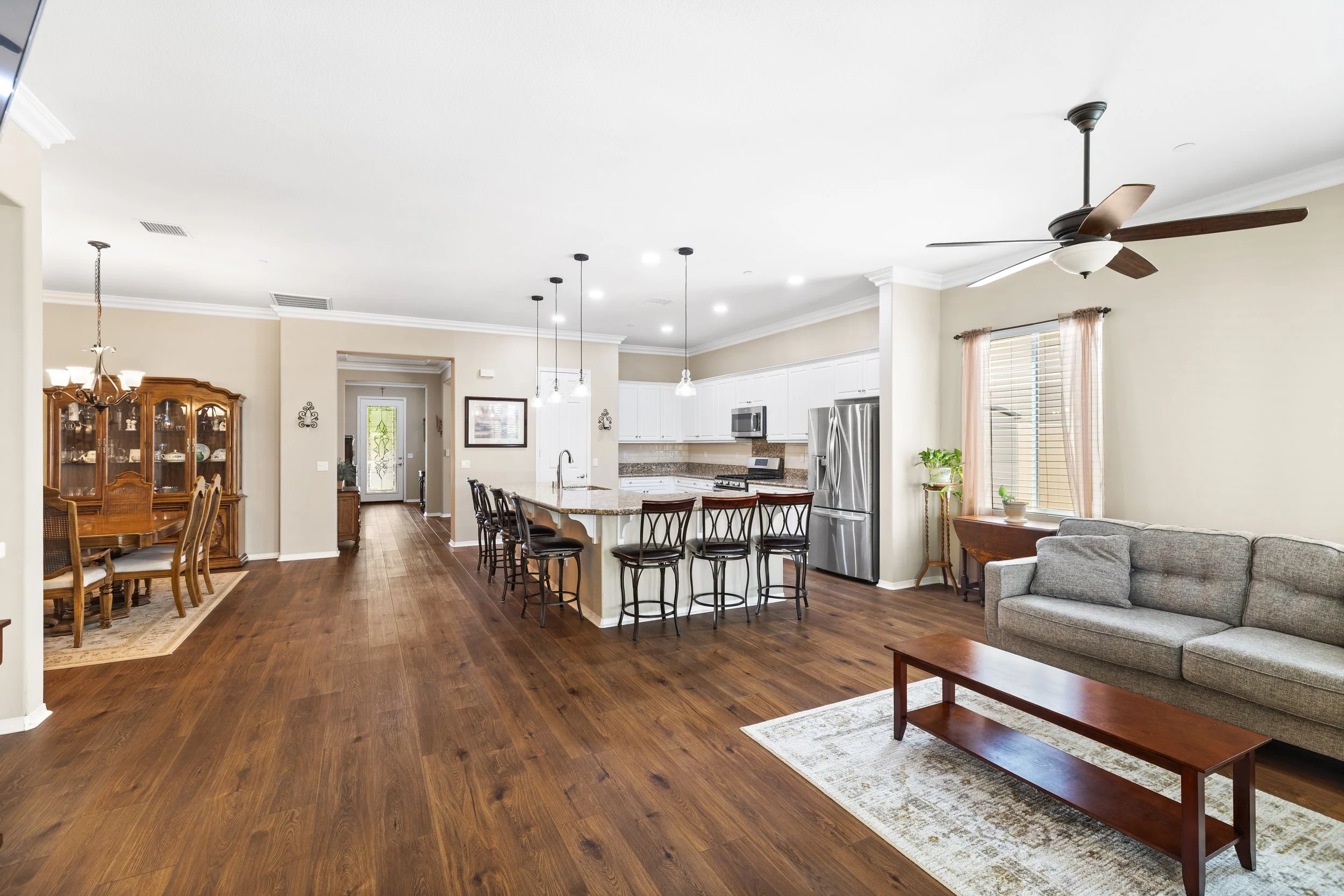 Open-concept living room and kitchen with hardwood floors, a gray sofa, a wooden coffee table, a ceiling fan, large windows, and white kitchen cabinets with a granite island and stainless steel appliances.