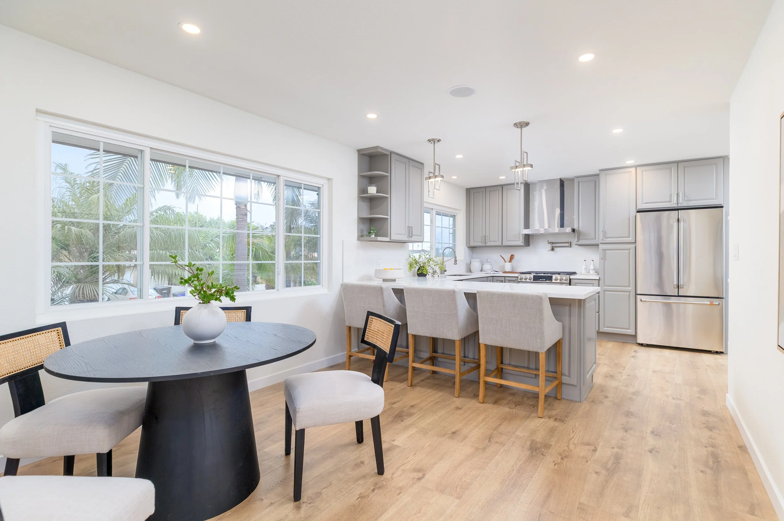 Bright, modern kitchen with light gray cabinets, stainless steel appliances, and a breakfast island with four chairs. A large window shows outdoor greenery. A dark wood dining table with chairs is nearby.