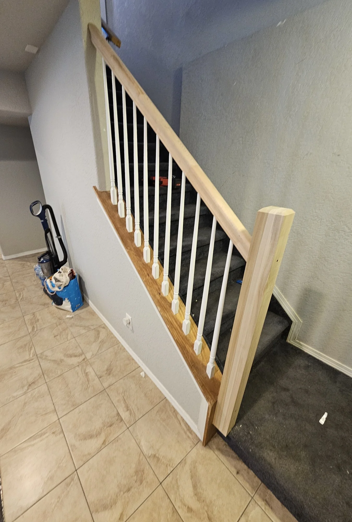 Staircase with an unfinished wood handrail, newel post, and white balusters beside a tiled hallway floor and carpeted stairs
