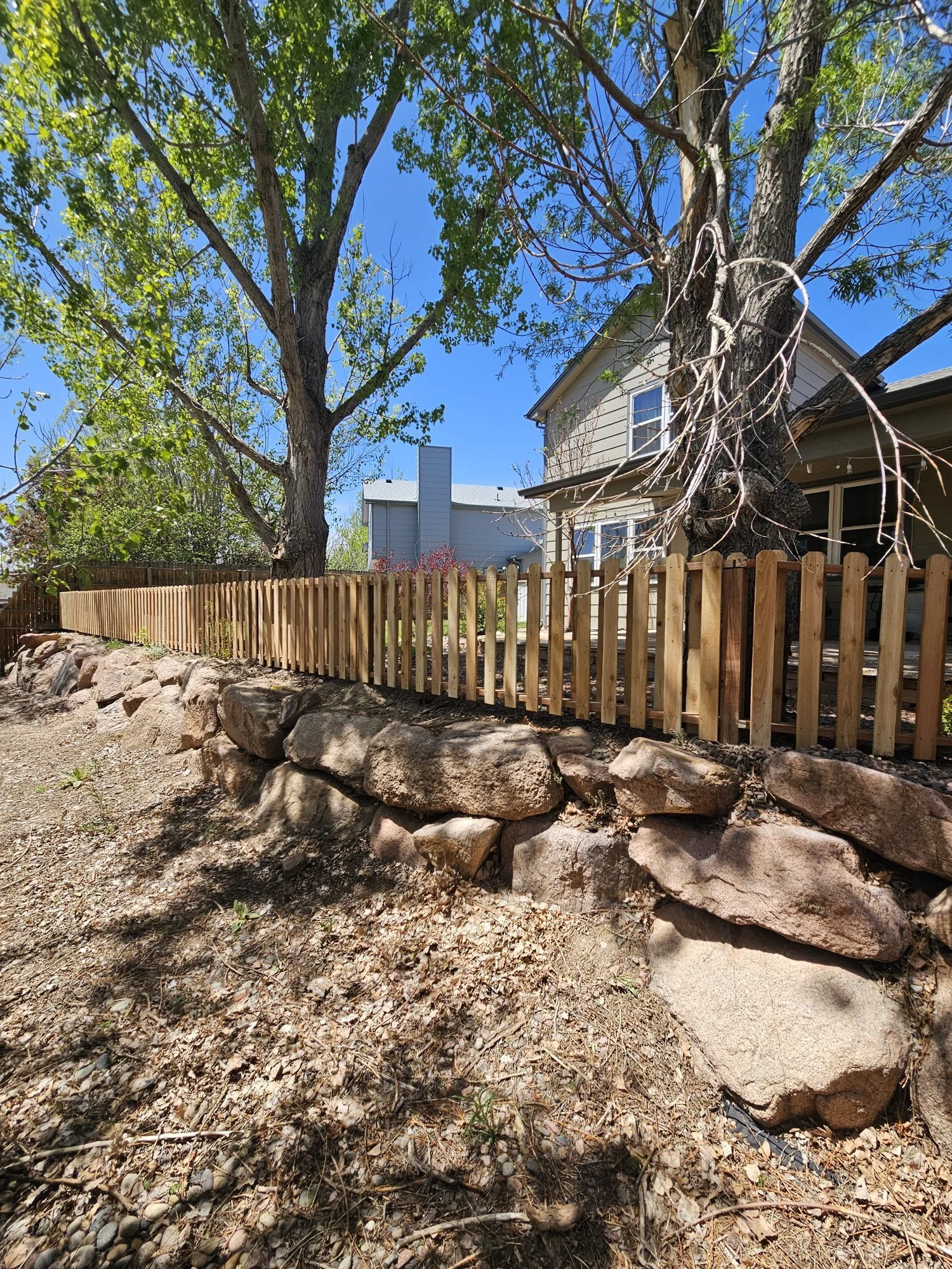New wood picket fence running along the top of a stacked boulder retaining wall in a backyard, with large trees and a house behind it