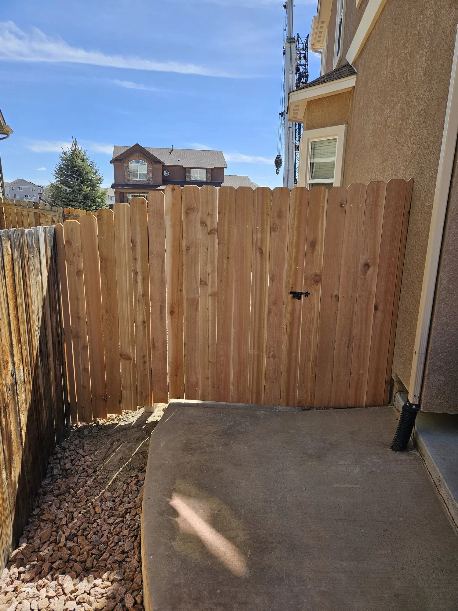 Wood privacy fence and gate with a black latch, next to a concrete pad and a gravel strip beside a tan stucco house