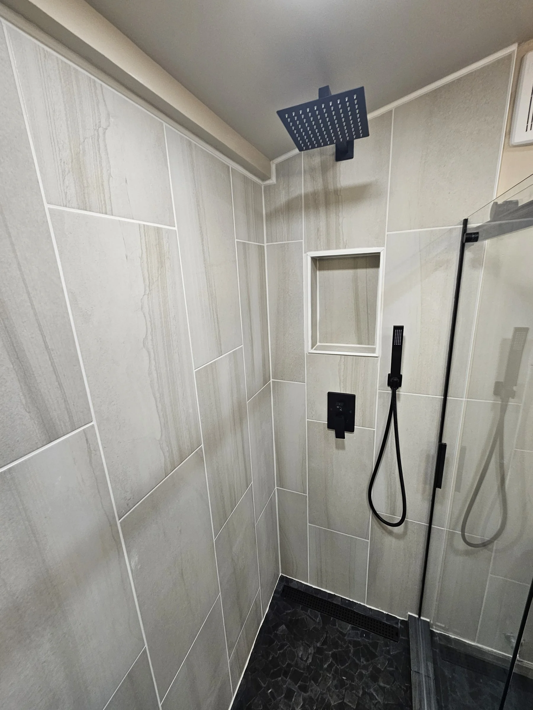 Walk-in shower with large light gray wall tiles, a recessed shelf niche, a matte black rain showerhead and handheld sprayer, black pebble tile floor with a linear drain, and a glass shower panel