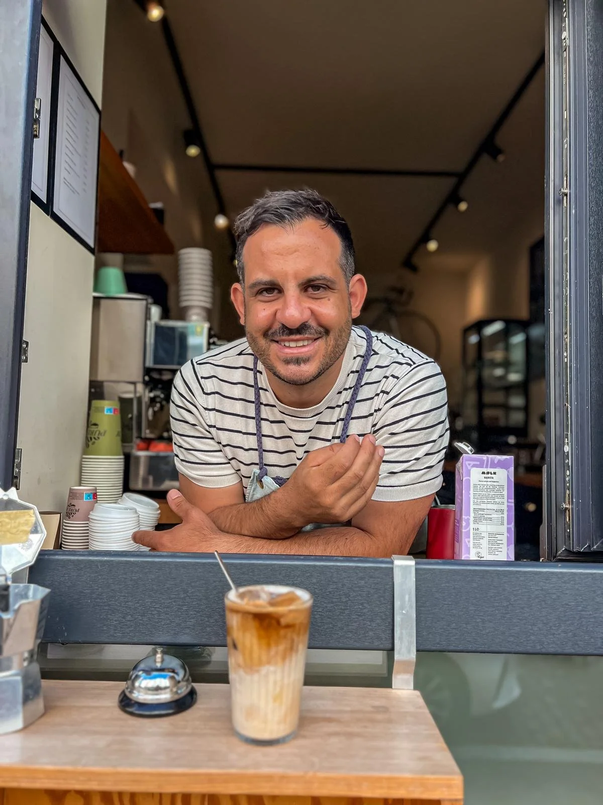 Massimo lächelt hinter der Cafétheke von VIA DEL WIR, davor steht ein Latte Macchiato.