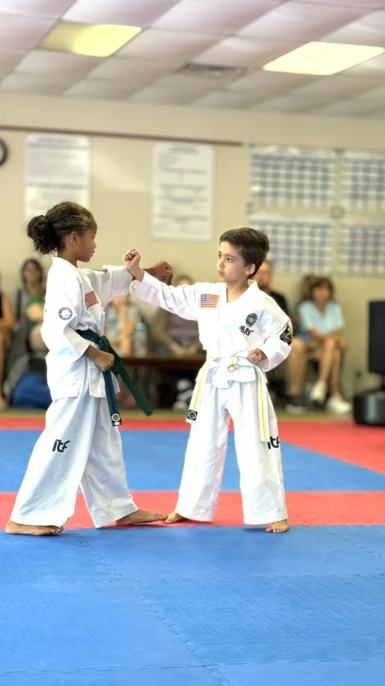 Two young children practicing martial arts in a dojo, one girl and one boy, wearing white gi uniforms with patches and belts, exchanging a handshake on a blue and red mat with spectators seated in the background.