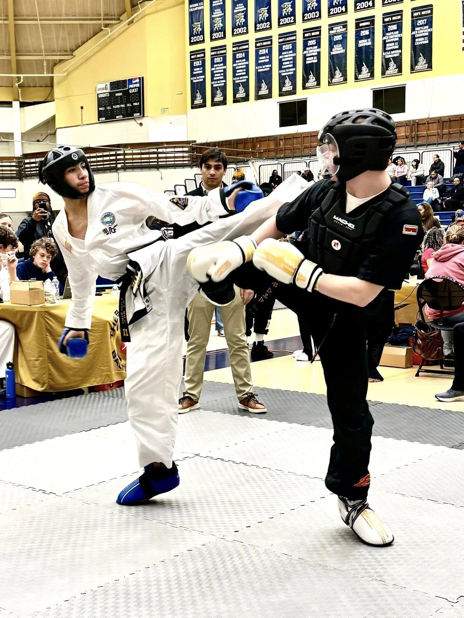 Two martial artists sparring in taekwondo gear on a mat, with spectators and banners in the background.