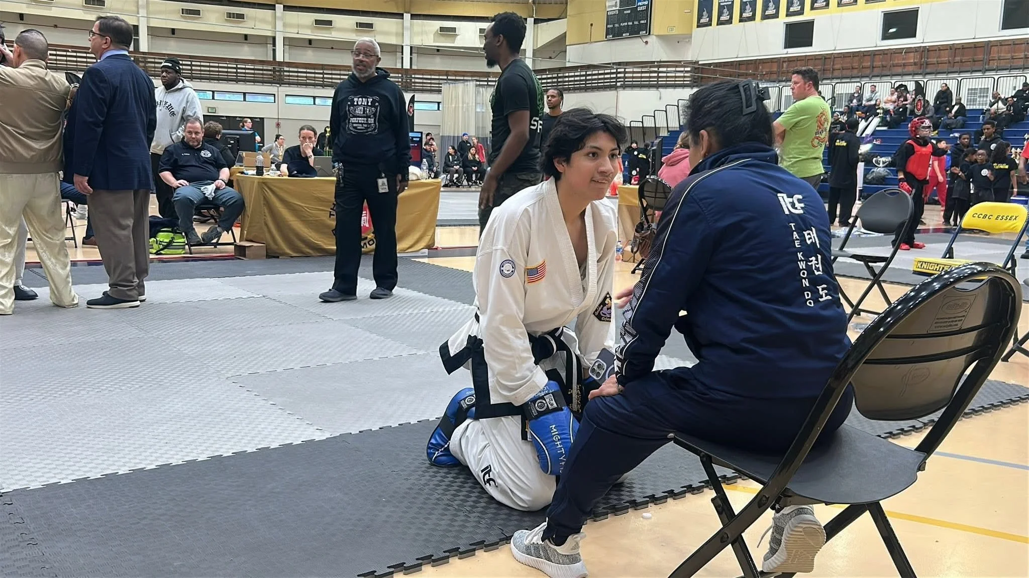A woman in a white martial arts uniform kneeling on the mat, talking to a seated woman in a navy jacket at a martial arts tournament in a gymnasium filled with people and spectators.