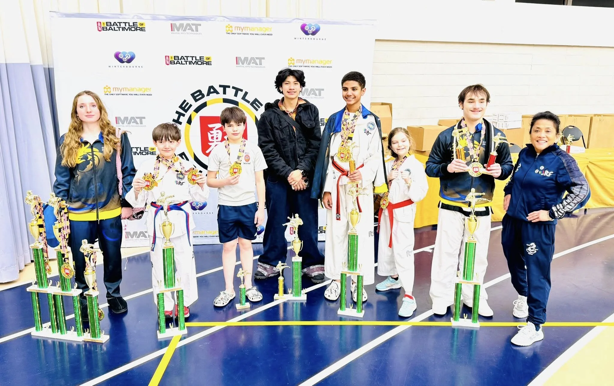 Group of children and two adults at a martial arts competition medal ceremony, standing with trophies on a gymnasium floor, with a banner and boxes in the background.