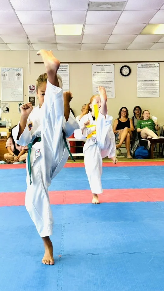 Two children practicing karate kicks in a martial arts dojo with spectators watching in the background.