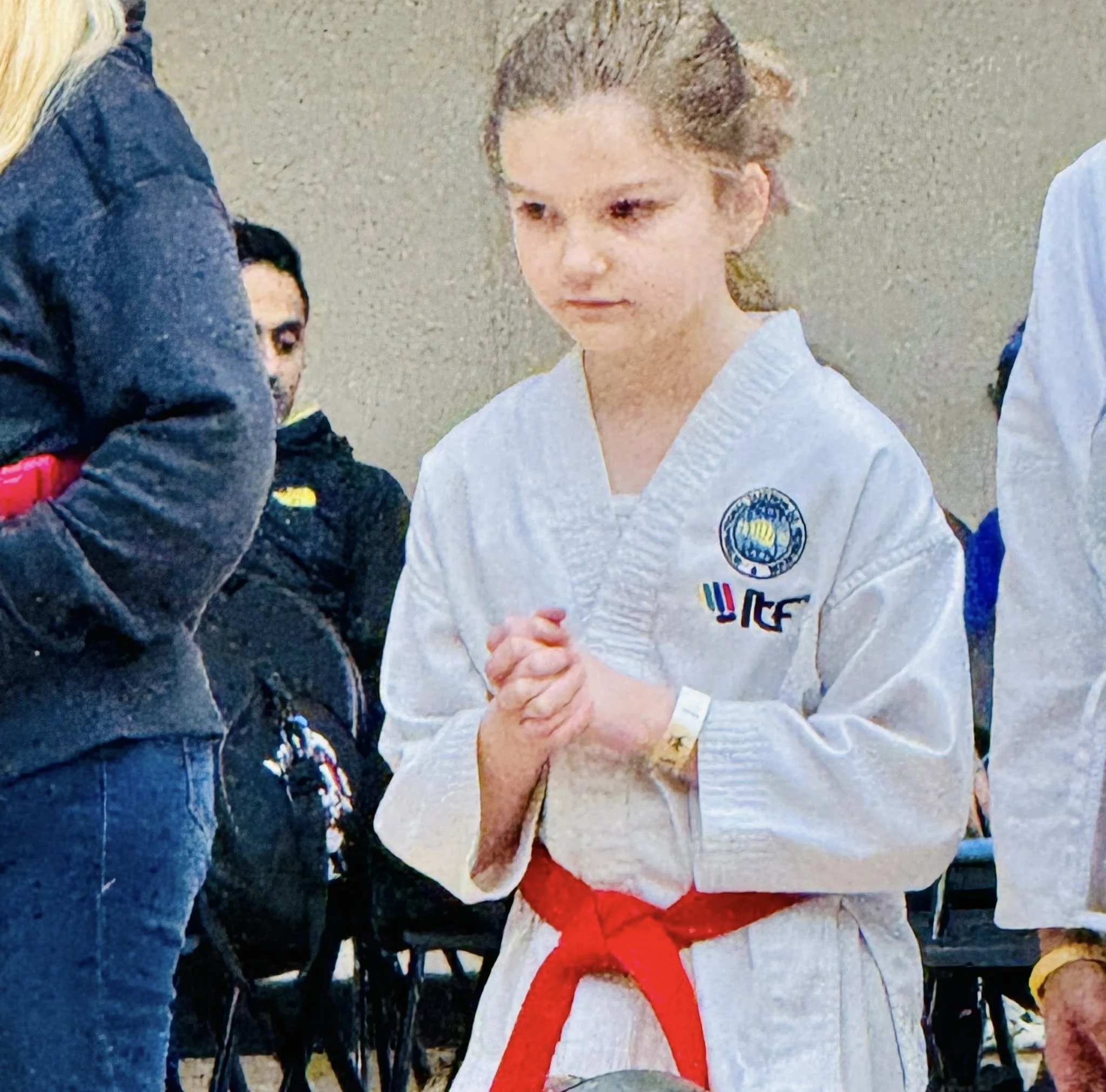 A young girl in a white martial arts uniform with a red belt, standing with her hands clasped, appears to be in a martial arts class or competition.