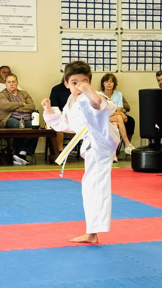 Young boy wearing a white martial arts uniform performing a high kick in a dojo with a red and blue mat, with spectators seated in the background.