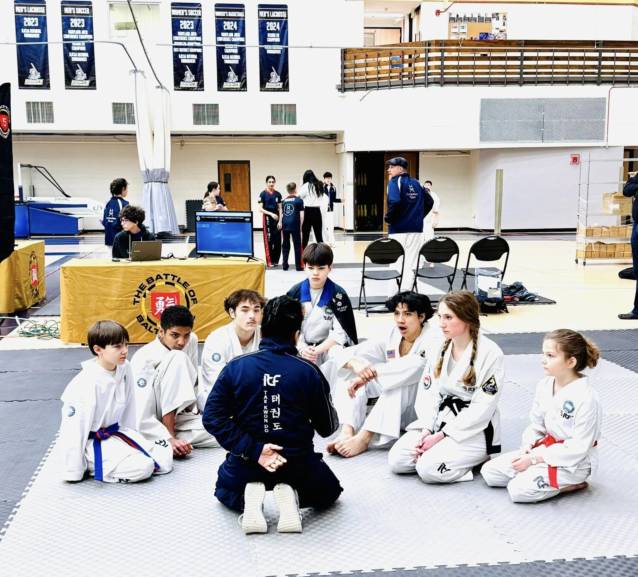 Martial arts students, including boys and girls, sitting on the mat listening to their instructor during training in a gymnasium with banners hanging from the wall.