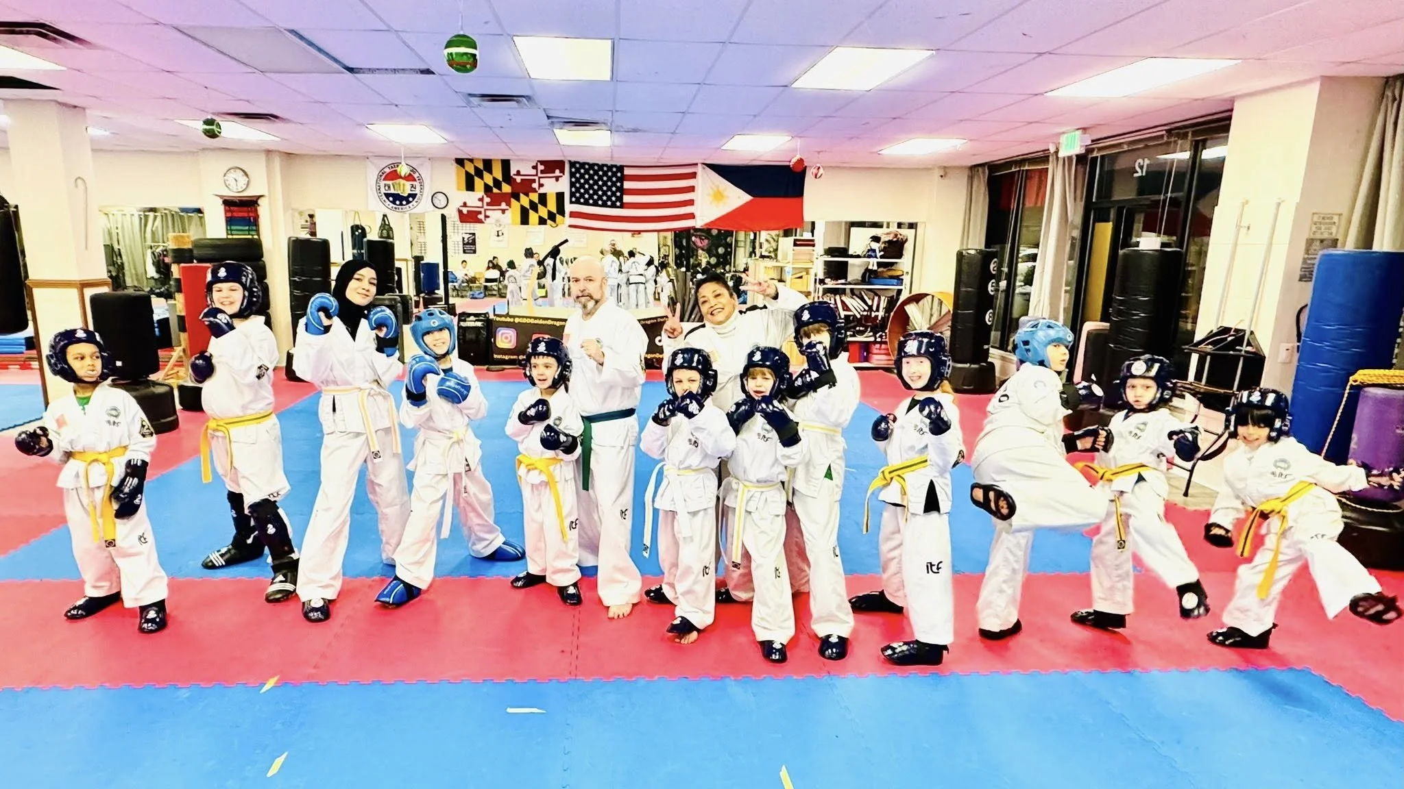 Group of children and instructors in a martial arts dojo, all wearing Brazil and USA flags in the background, practicing martial arts in white uniforms with colored belts, some wearing blue helmets, in a well-equipped martial arts gym.