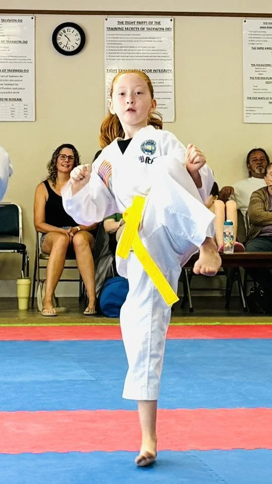 Young girl in a martial arts uniform performing a kick in a martial arts studio with spectators in the background.