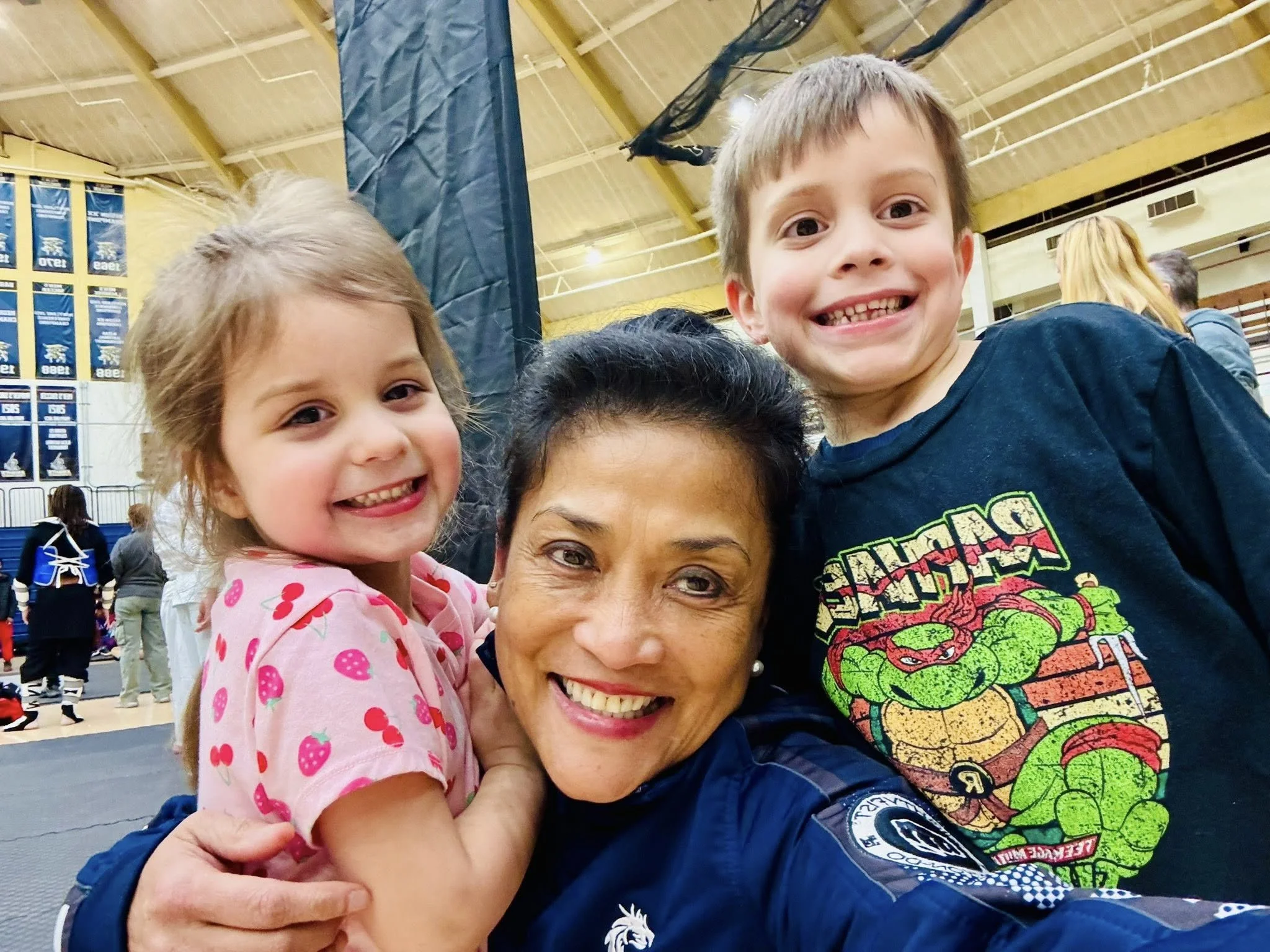 A woman in a police uniform hugging two smiling children in a gymnasium. The girl on the left is wearing a pink shirt with strawberries, and the boy on the right is wearing a Teenage Mutant Ninja Turtles shirt.