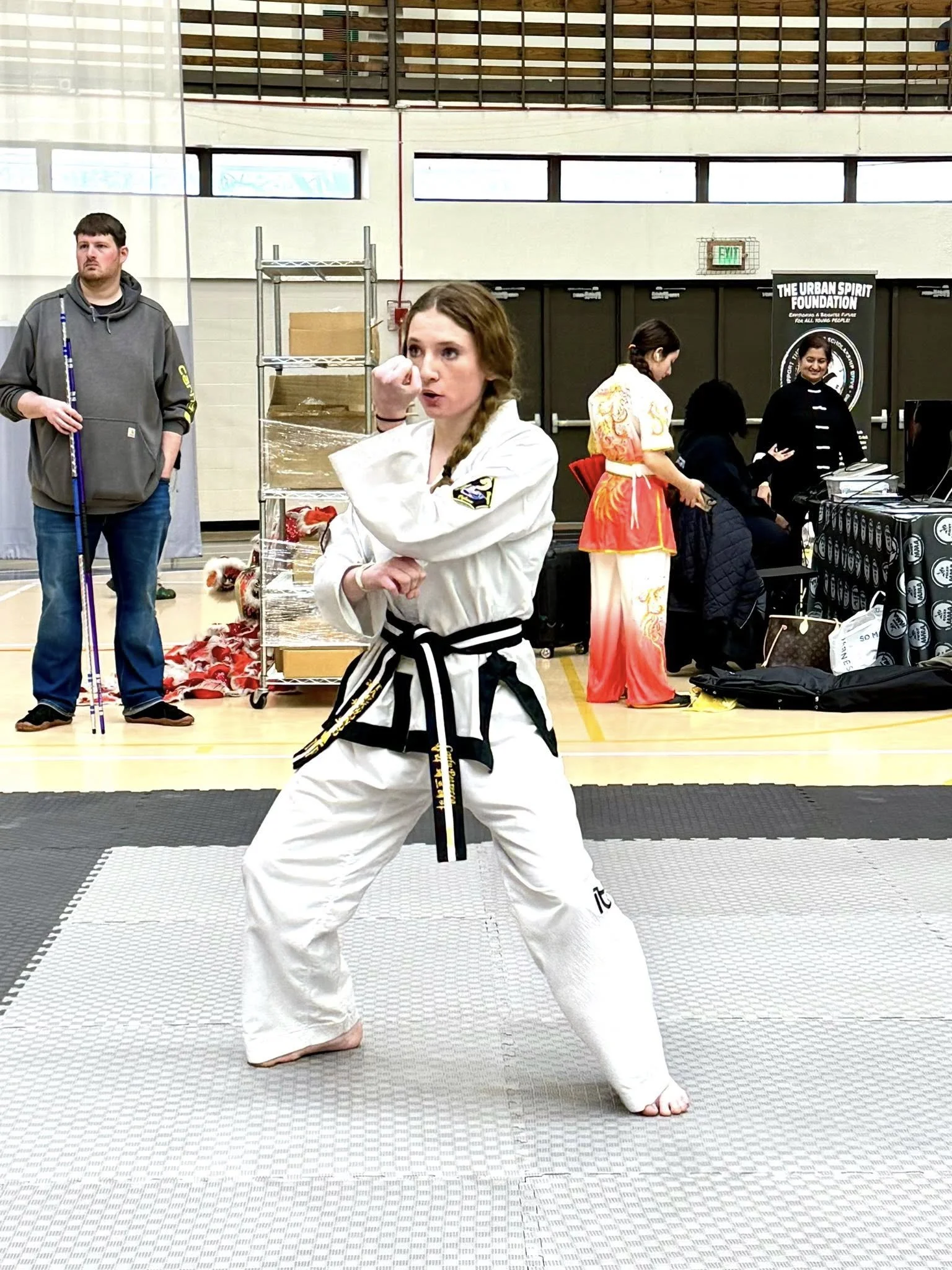 A female martial artist in a white gi with a black belt practicing a pose on a gray mat in an indoor gym, with people and booths in the background.