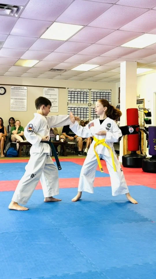 Two children in martial arts uniforms practicing a sparring match on a blue and red mat in a dojo, with an audience in the background.