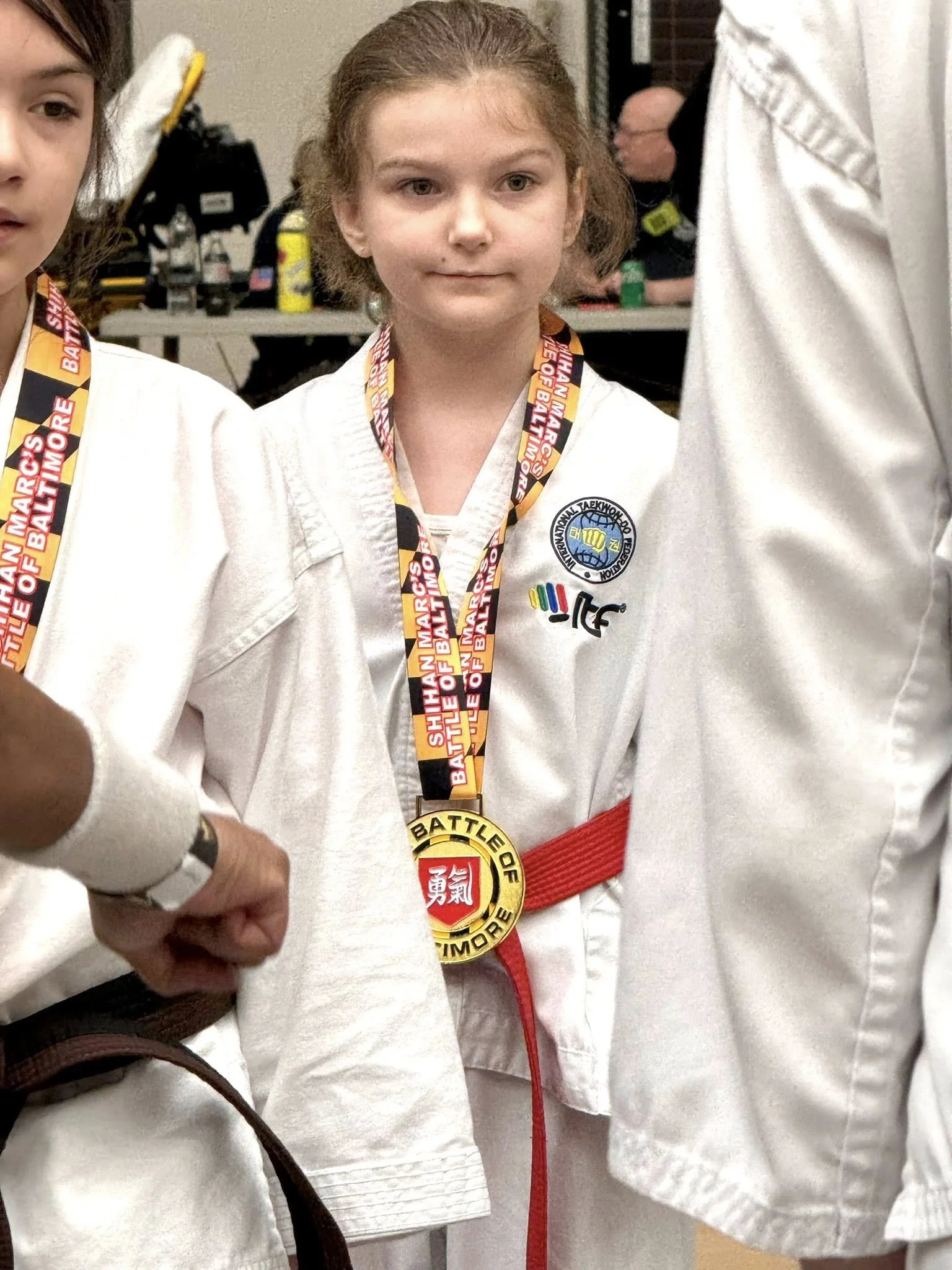 Young girl in a white martial arts uniform with a red belt, wearing medal around her neck, standing among other martial artists at a competition or event.