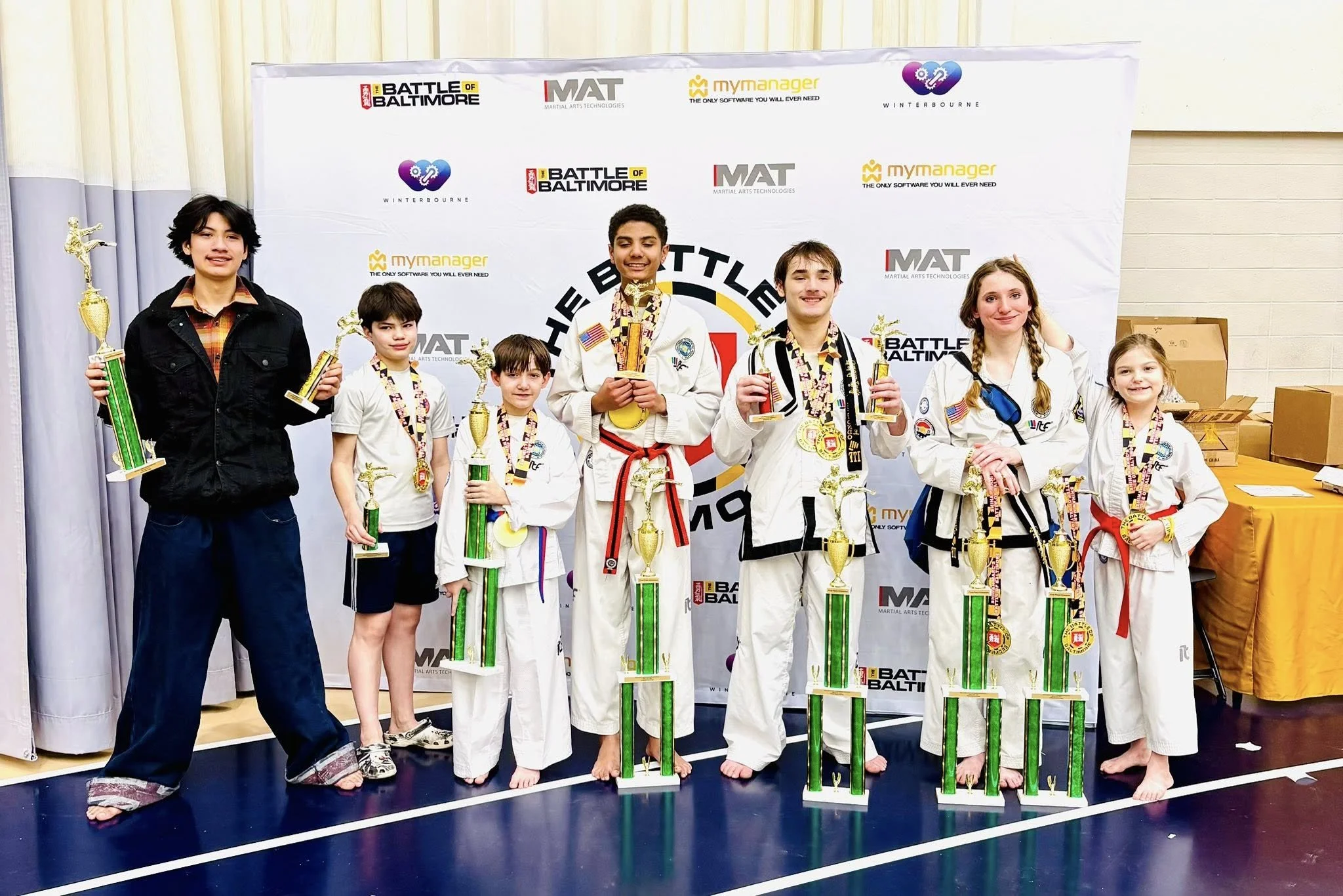 Group of young martial artists in uniforms holding trophies and medals, standing on a podium after a competition, with a backdrop displaying event logos.