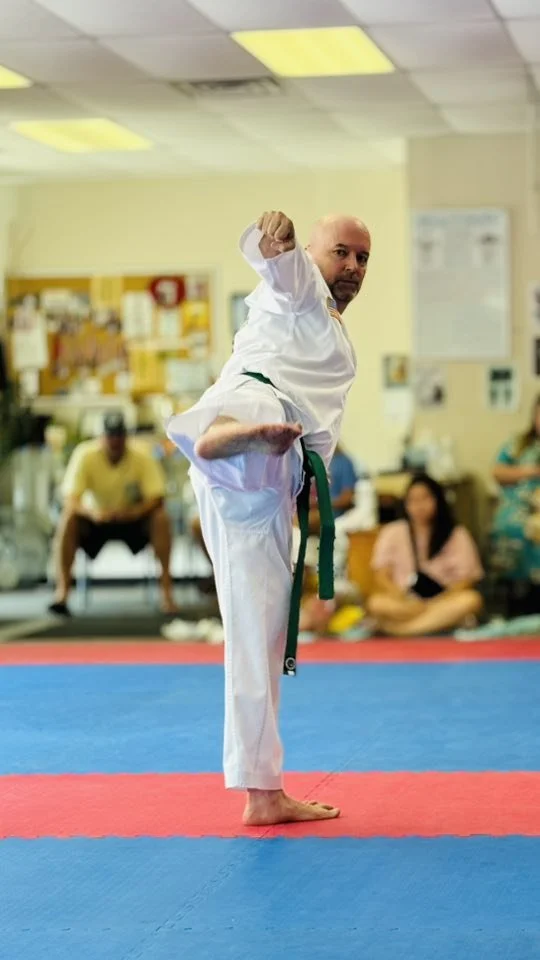 Martial artist performing a high kick in a dojo or training center, with spectators seated in the background.