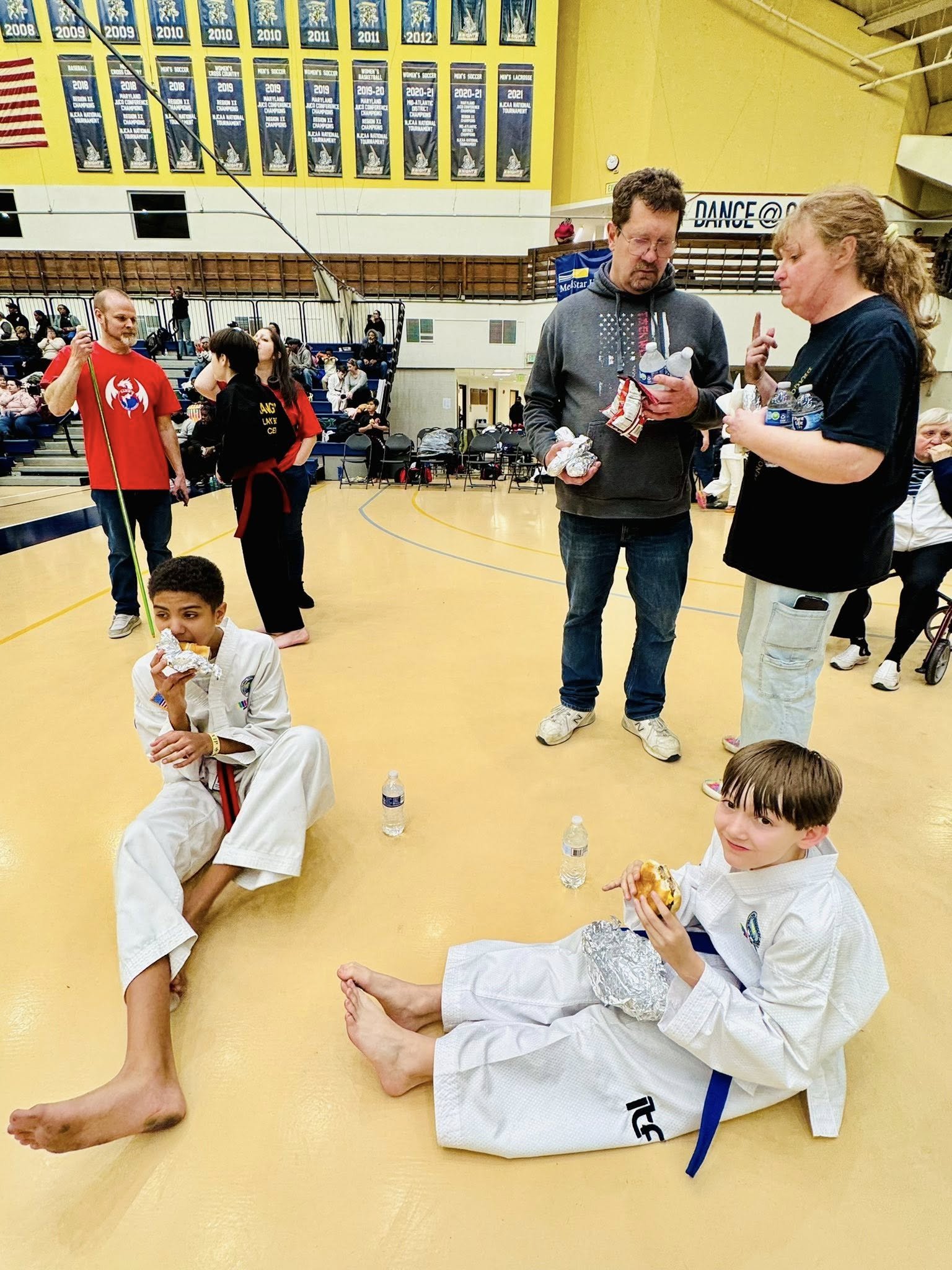 Two children in martial arts uniforms sitting on the floor in a gymnasium, eating food wrapped in foil, with water bottles beside them. The gym has banners hanging on the wall and people in the background, some standing and some sitting on bleachers.