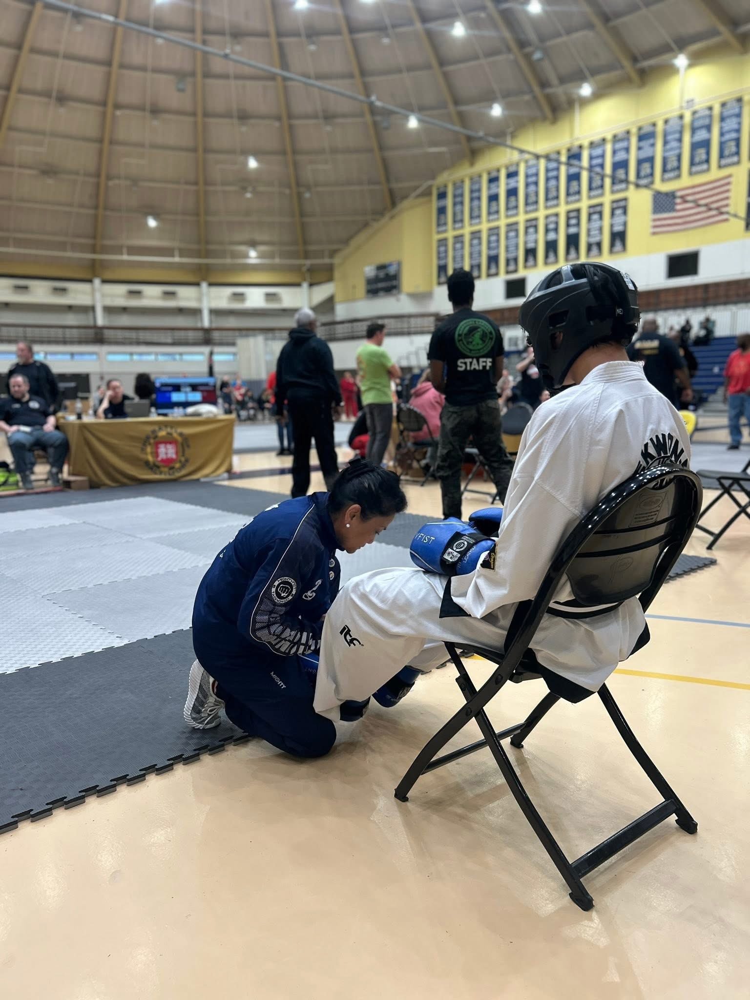 Martial arts event inside a gymnasium with a fighter in a white uniform sitting on a chair, wearing a helmet and gloves, while a woman in a blue uniform adjusts his foot.