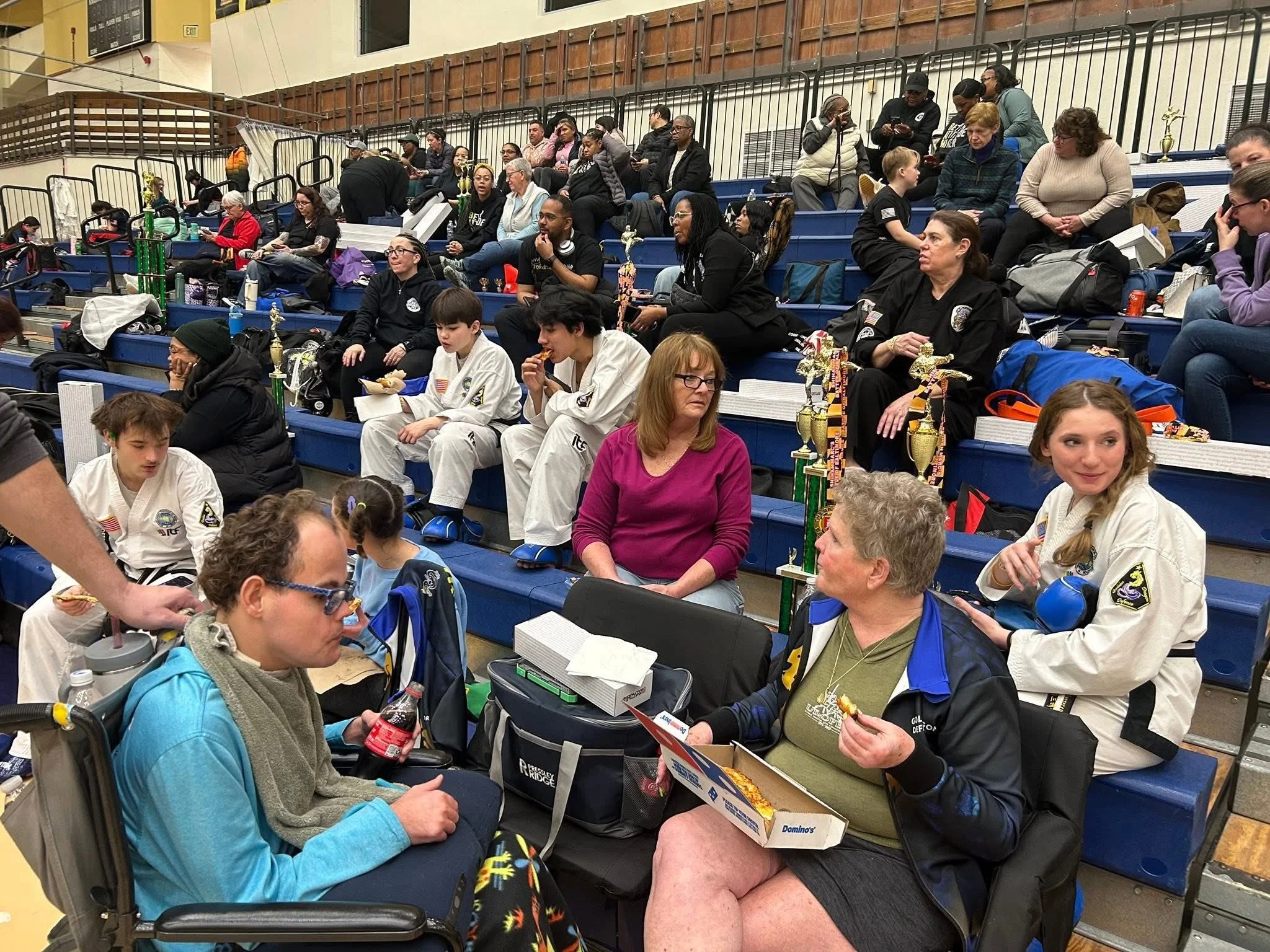 People seated on bleachers inside a gymnasium at a martial arts tournament, with several trophies and medals displayed.