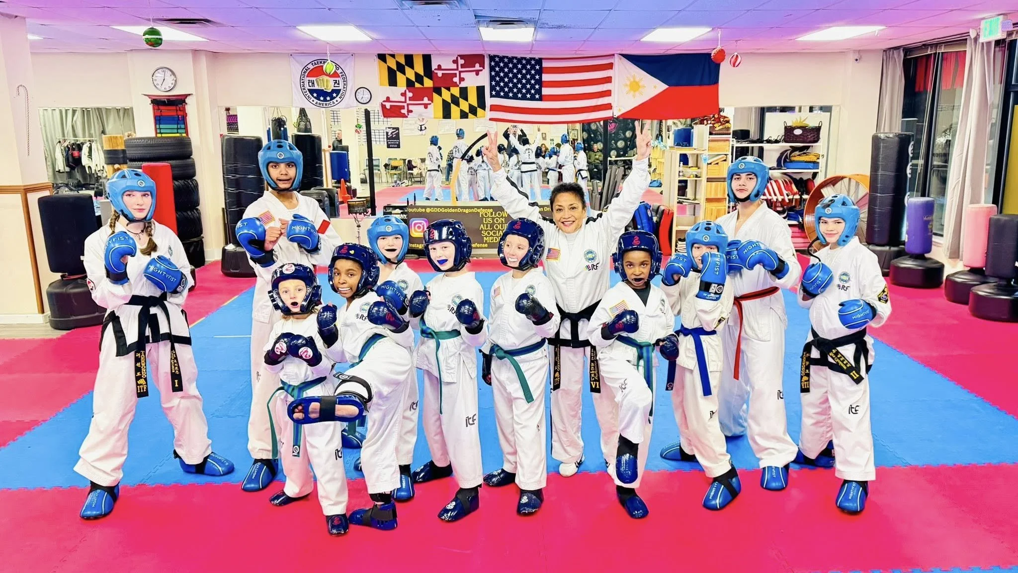 Group of children in taekwondo uniforms and helmets posing with a female instructor in a martial arts dojo, with flags hanging from the ceiling.