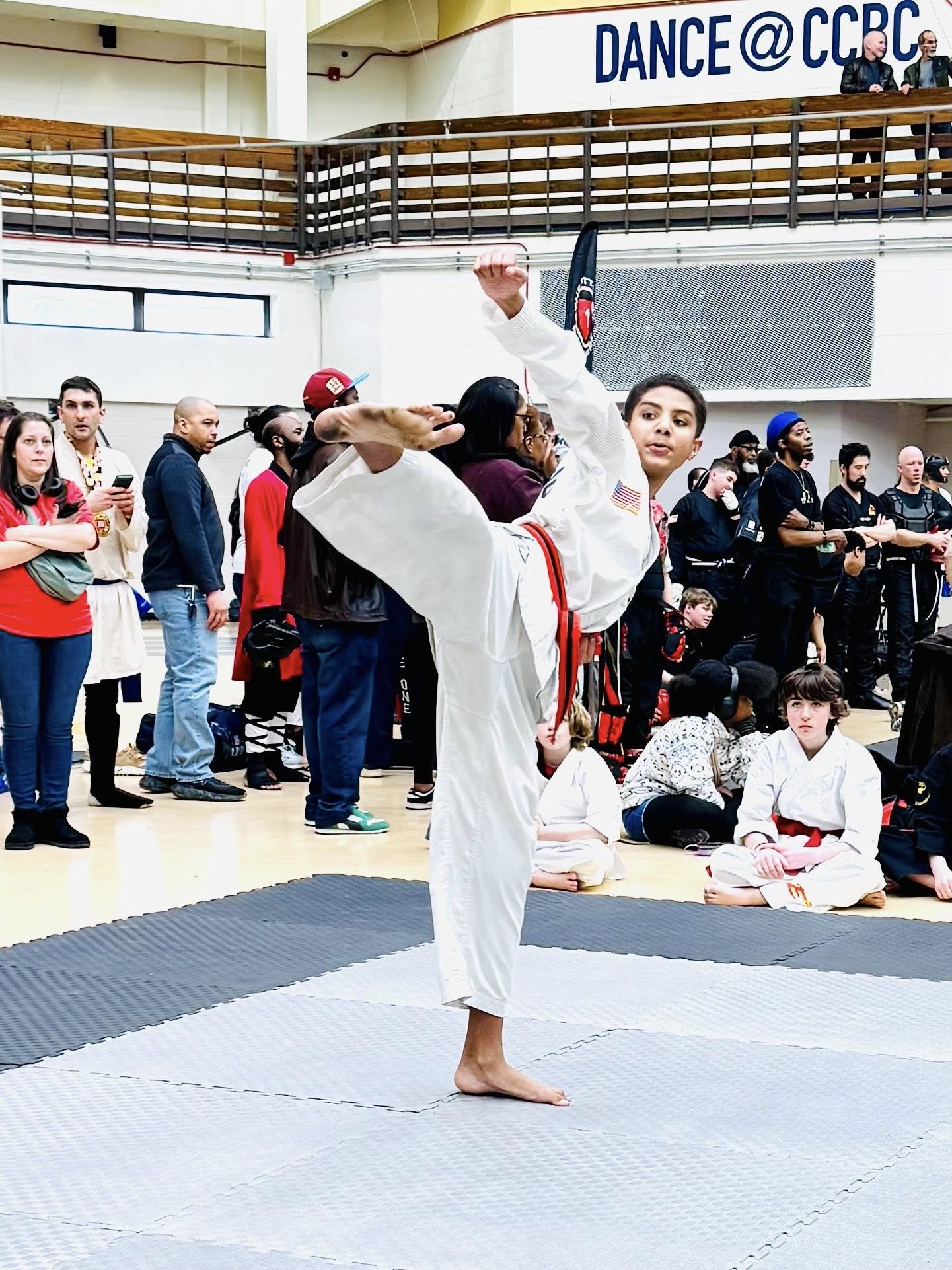 A young martial artist in a white gi performing a high side kick at a martial arts event, with a crowd of spectators and other competitors watching.