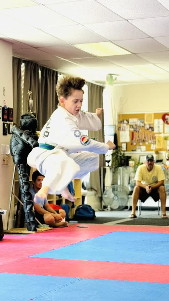 A young boy in a martial arts uniform is mid-air performing a jump in a martial arts dojo or classroom, with spectators and an instructor watching.