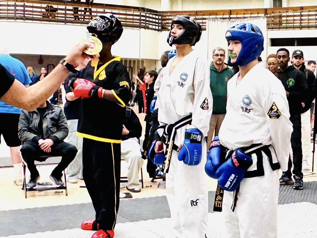 Three martial artists in white and black uniforms with protective helmets and gloves, receiving medals at a martial arts competition.
