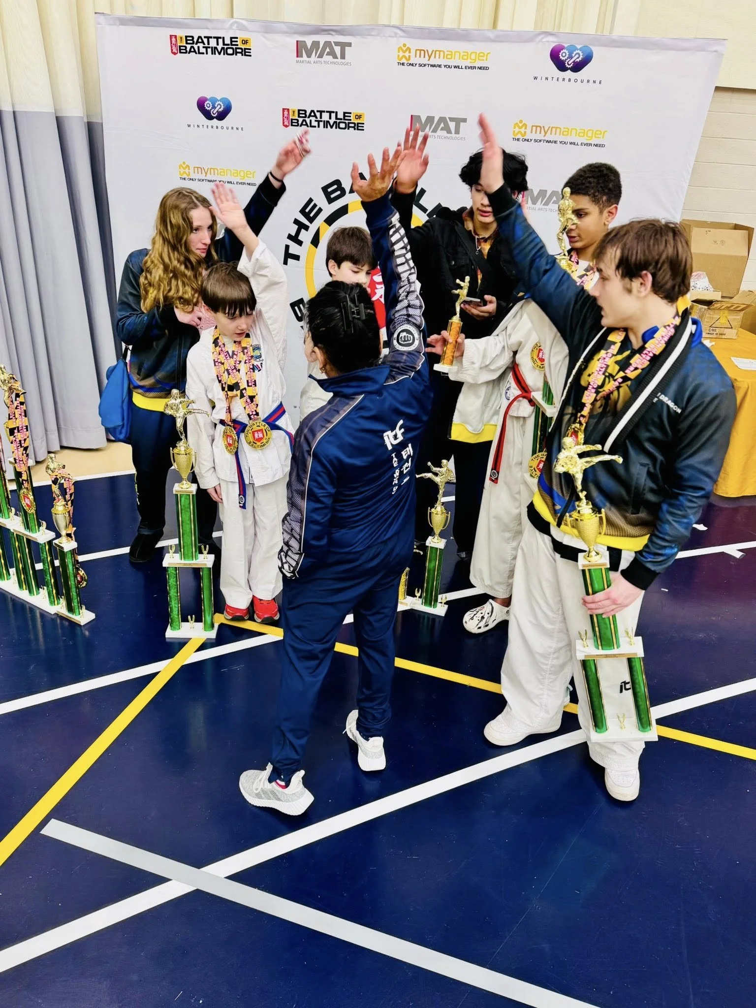 Group of children and adults celebrating at a martial arts competition, holding trophies, and high-fiving each other on a competition floor with a backdrop displaying multiple logos.
